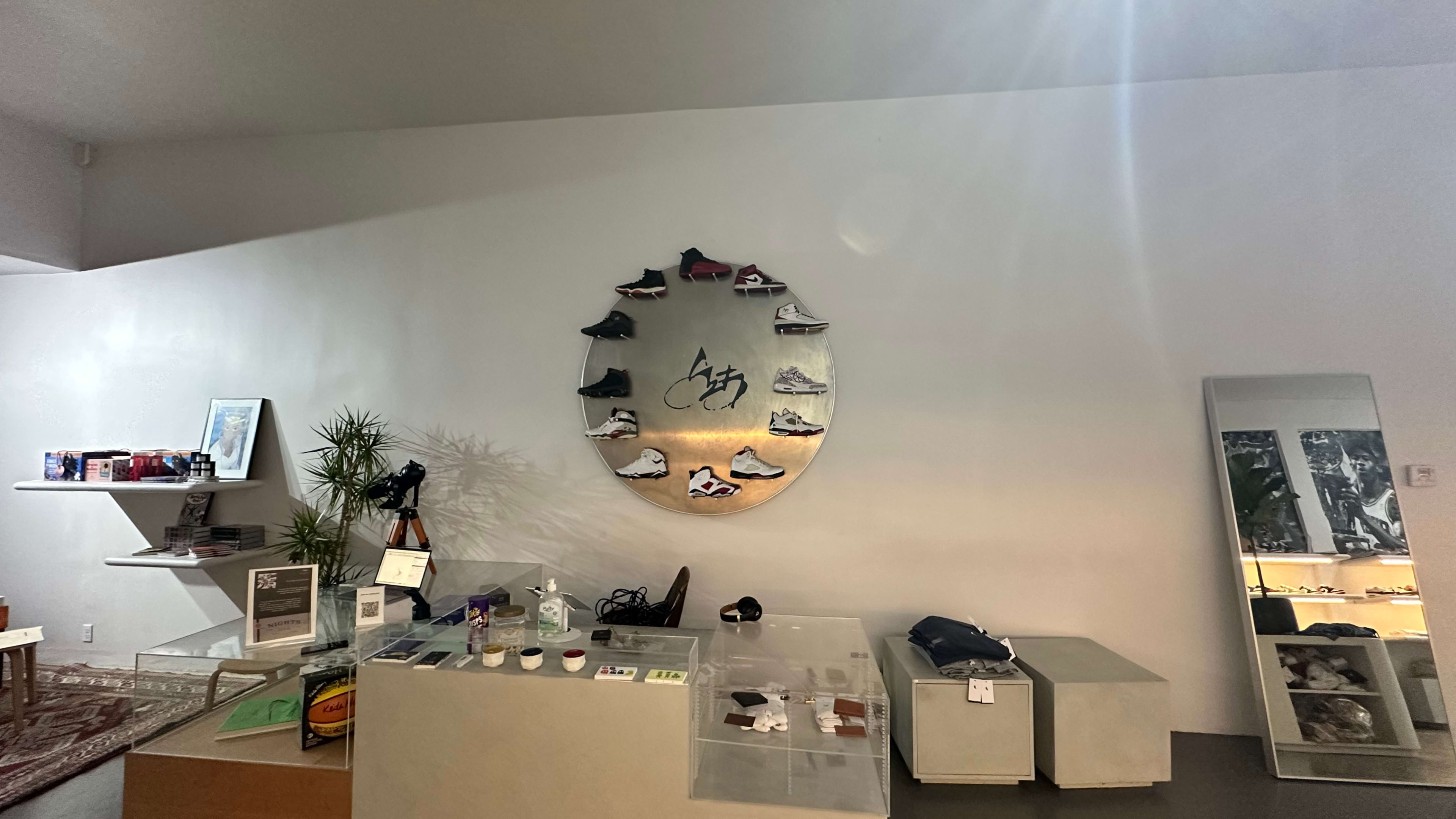 The image shows a minimalist retail space featuring a circular display of sneakers on the wall, along with various merchandise items on a counter and a mirror.
