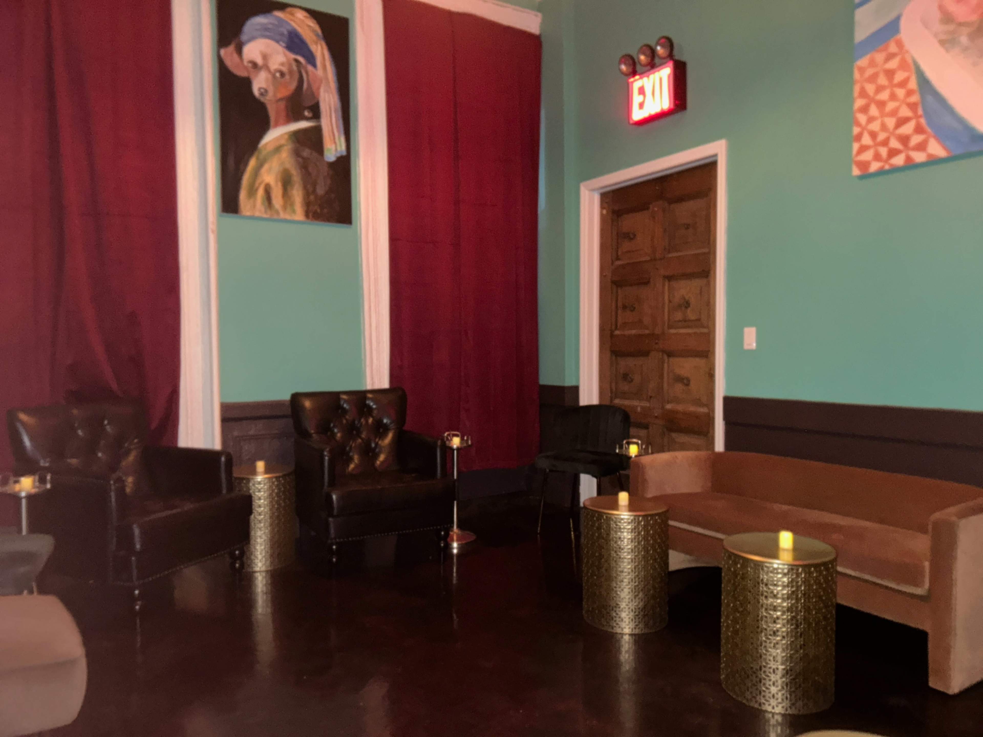 The image shows a seating area with two black armchairs and a brown sofa, surrounded by red curtains and adorned with a colorful painting on the wall.