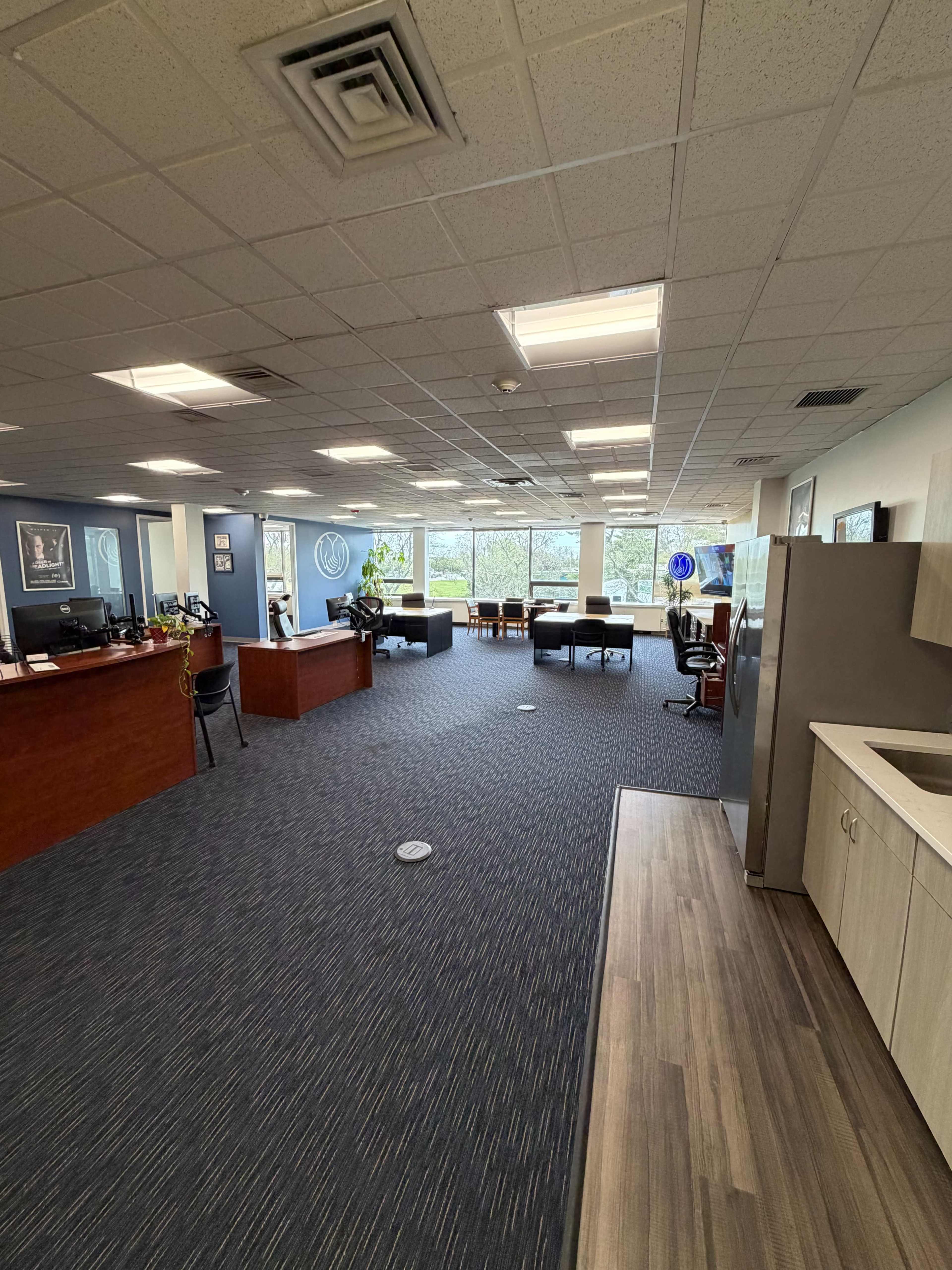 The image shows a spacious office area with workstations, a kitchenette, and large windows providing natural light.