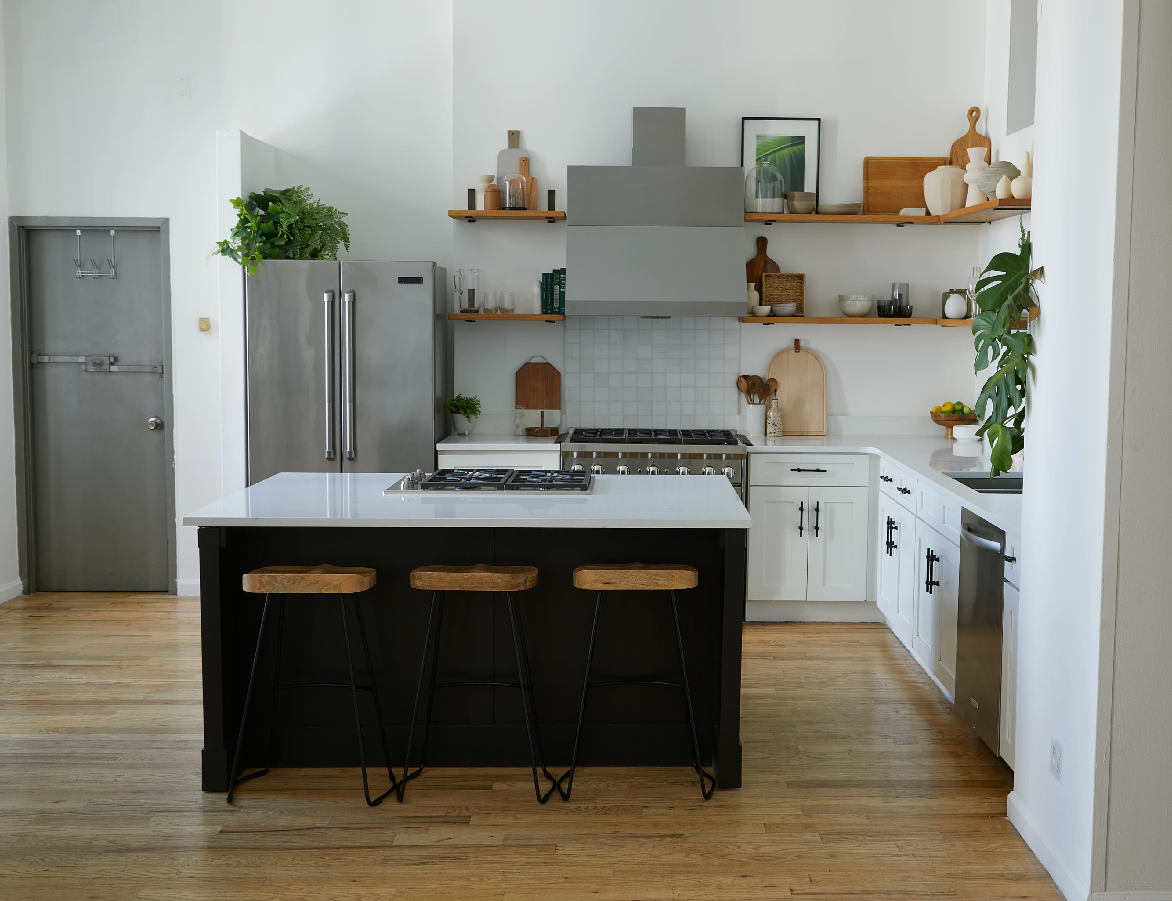 The image shows a modern kitchen featuring a central island with bar stools, stainless steel appliances, and open shelving displaying various kitchen items.