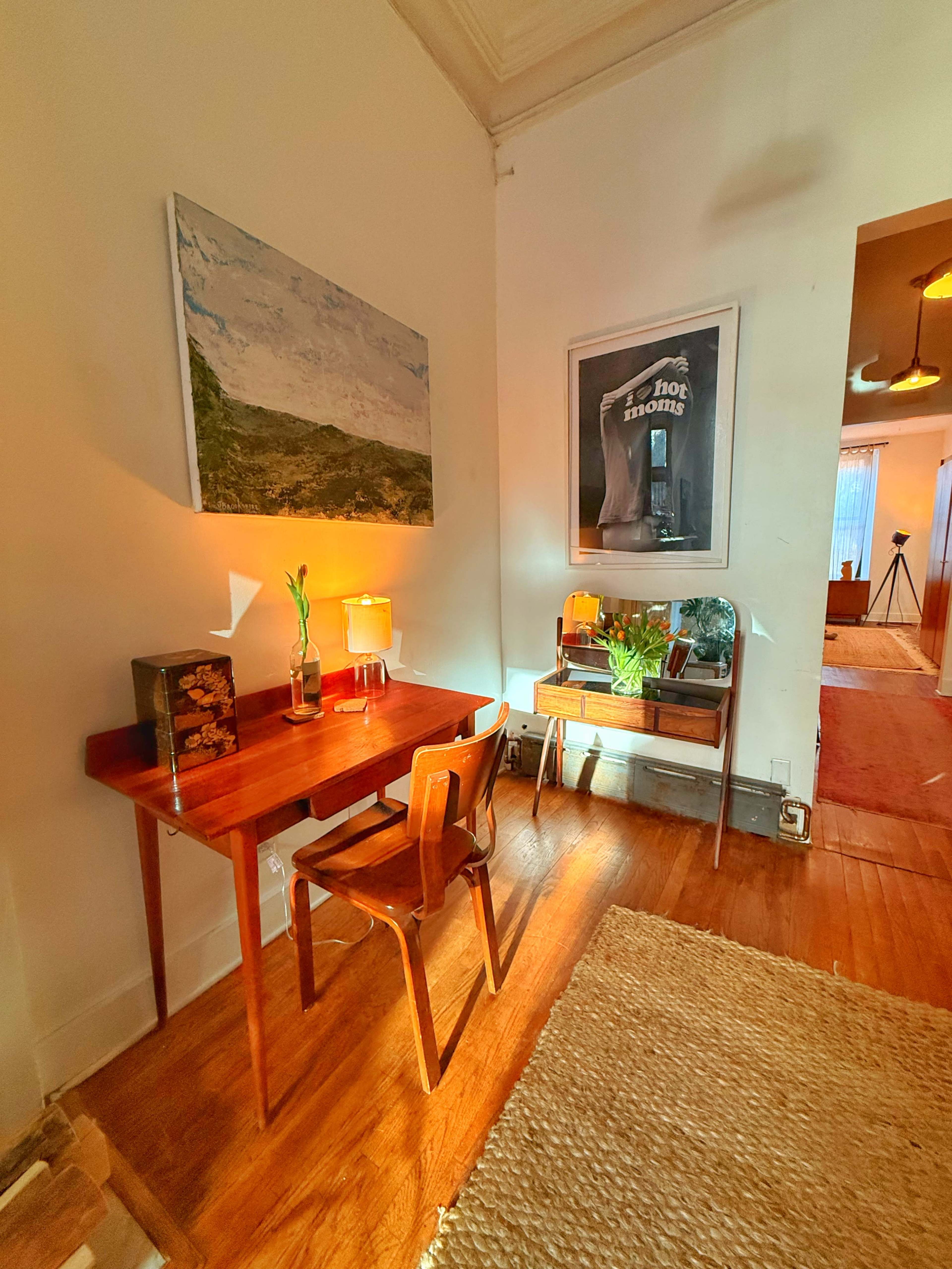 The image shows a well-lit corner of a room featuring a small wooden desk with a chair, a lamp, an artwork on the wall, and a reflective surface that includes a framed shirt.