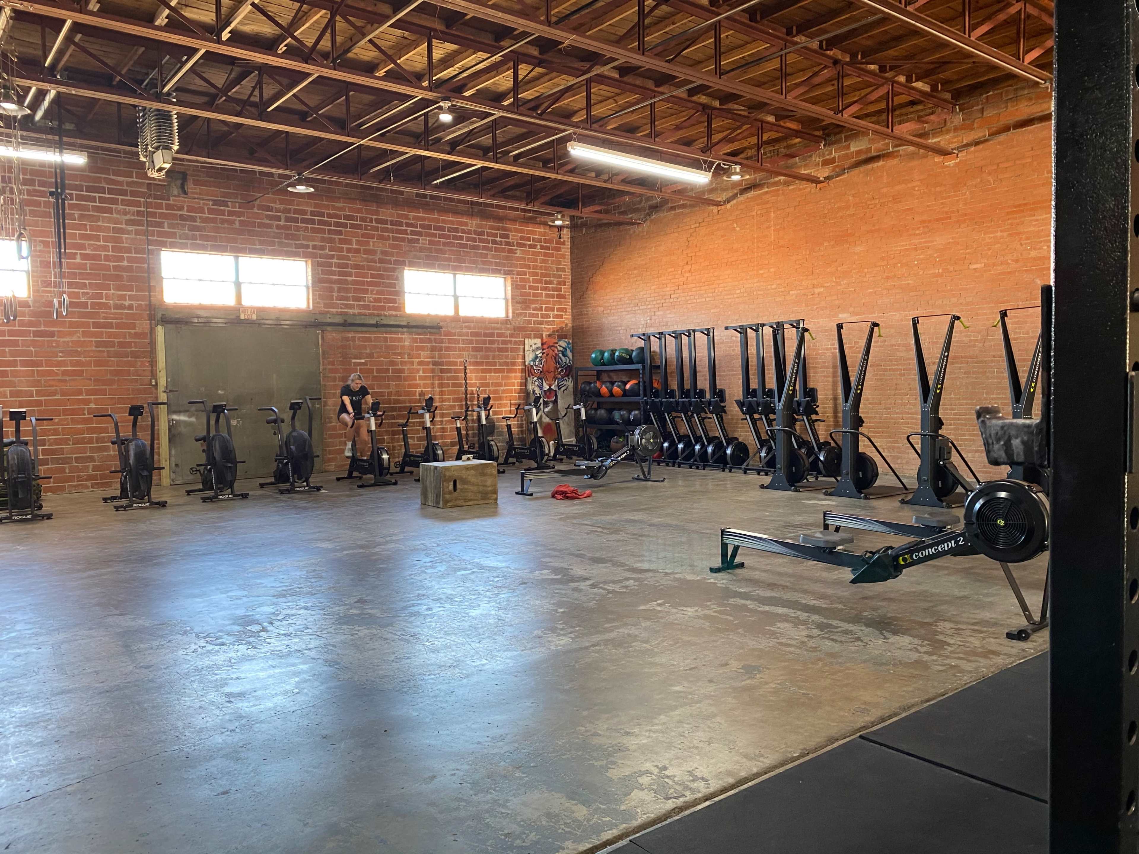 A spacious gym interior features exercise equipment, including rowing machines and air bikes, along with a wooden box and weights against the brick wall.