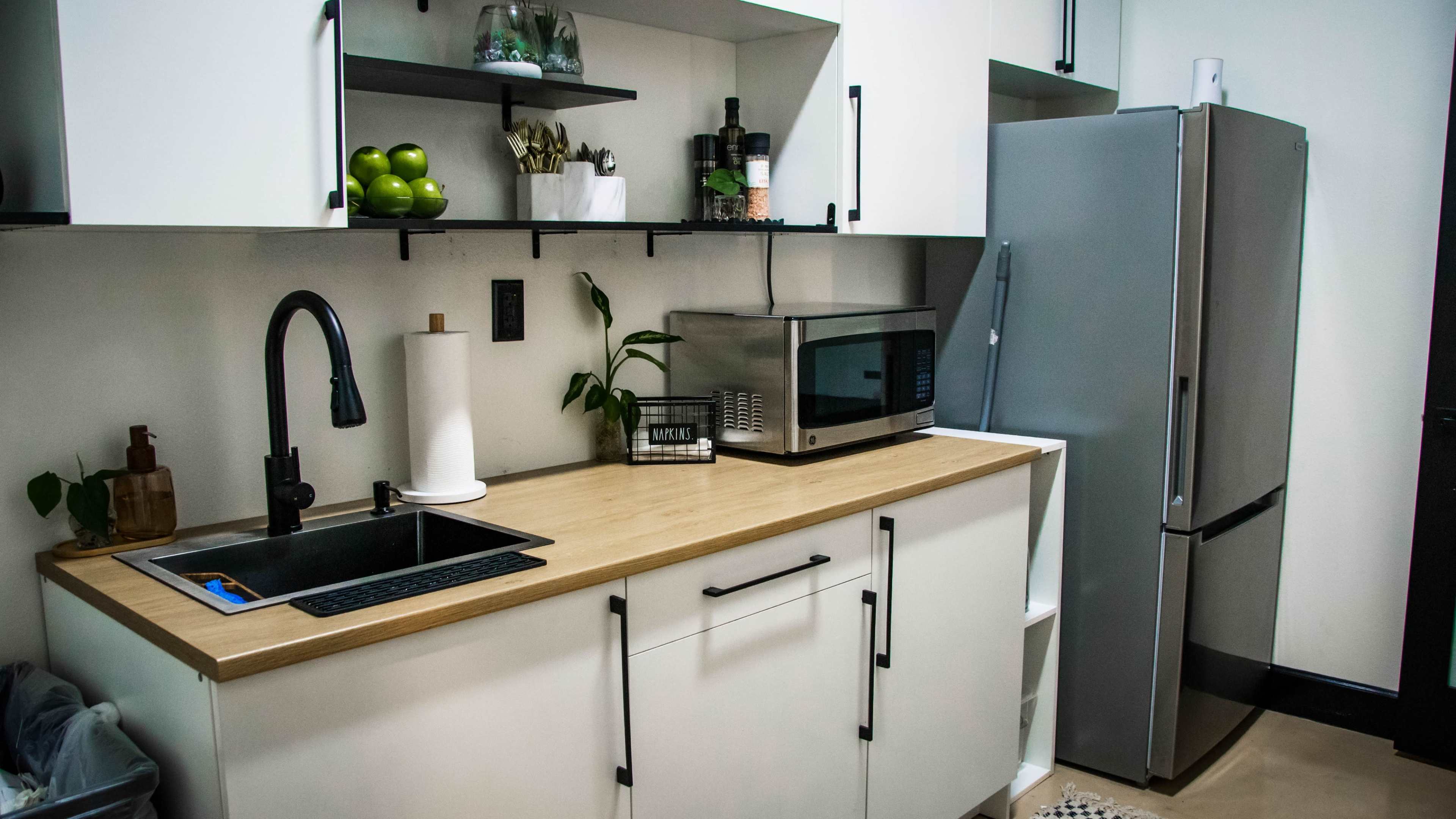 The image shows a modern kitchen with a wooden countertop, a sink with a black faucet, a microwave, and a stainless steel refrigerator.
