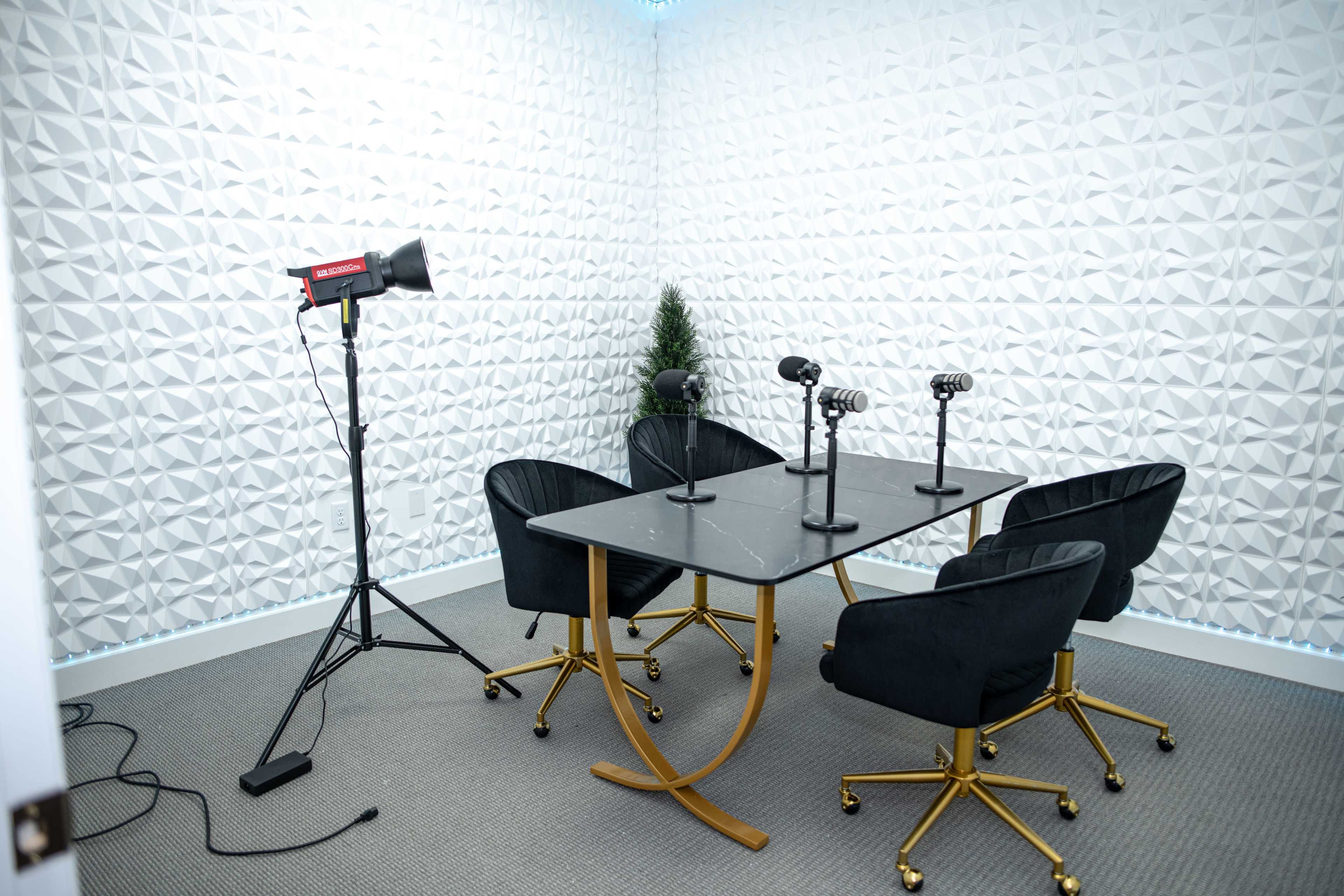 The image shows a small recording studio with a black table, four chairs, three microphones, and a spotlight, set against a textured white wall.