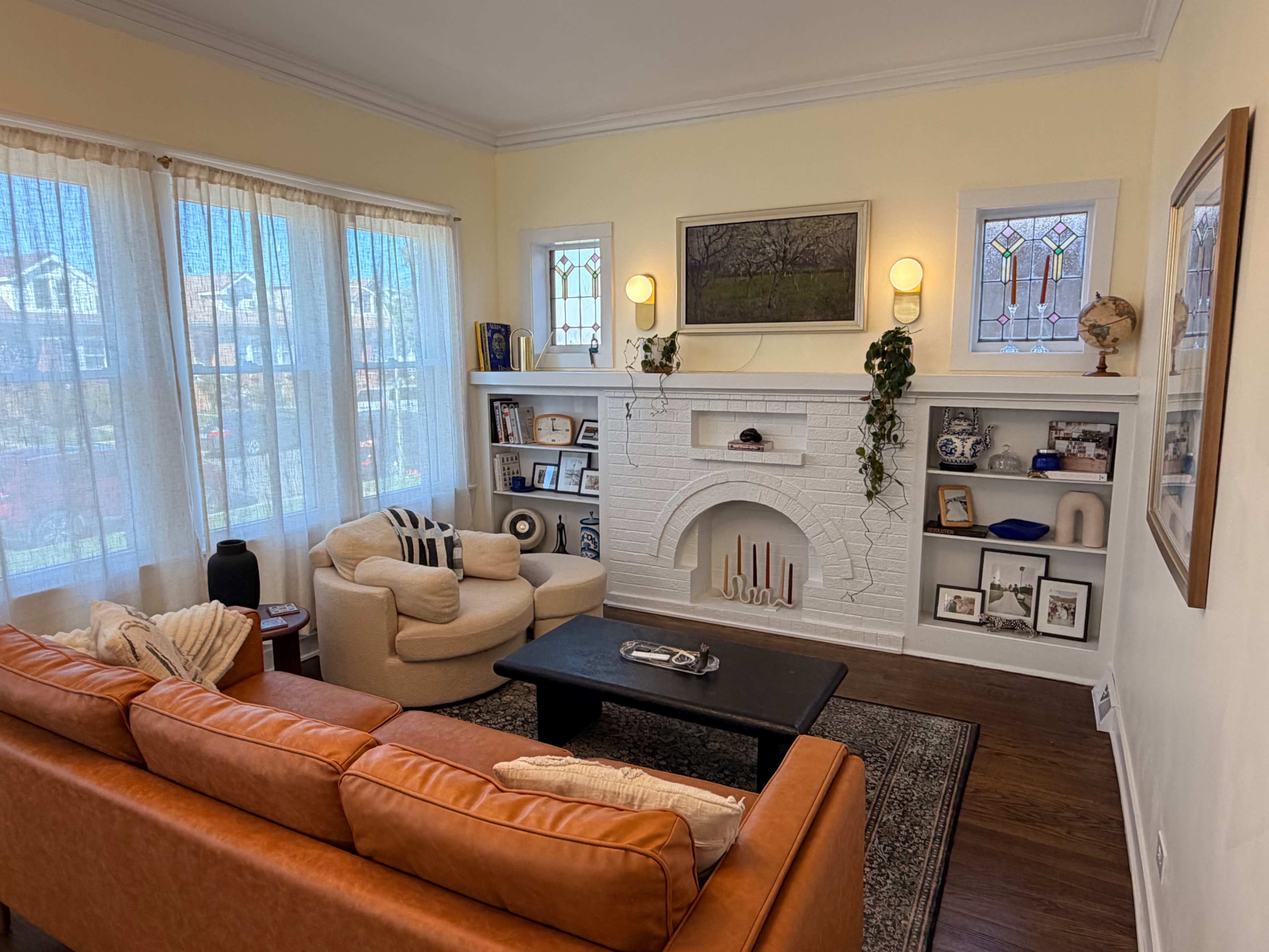 A living room features a light-colored sofa, a round armchair, a black coffee table, and shelves displaying decorative items against a backdrop of large windows and stained glass.