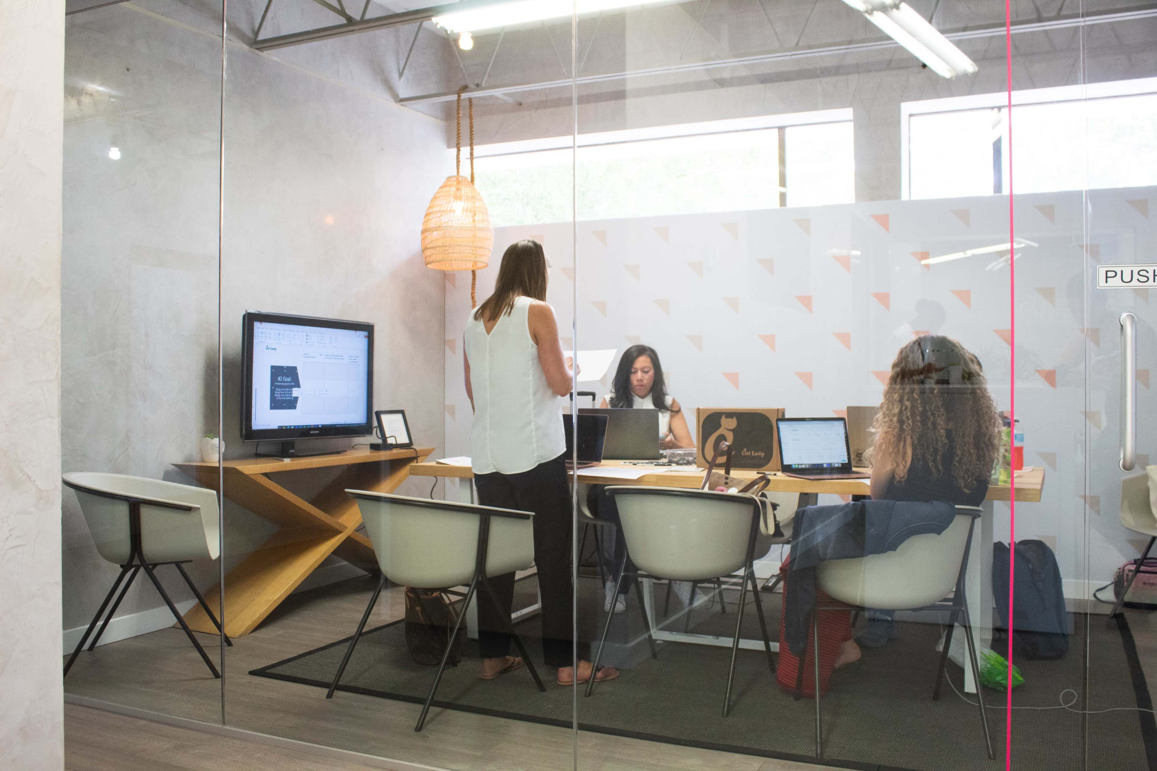 Three people are working at a conference table in a modern office space, with a large screen displaying information in the background.