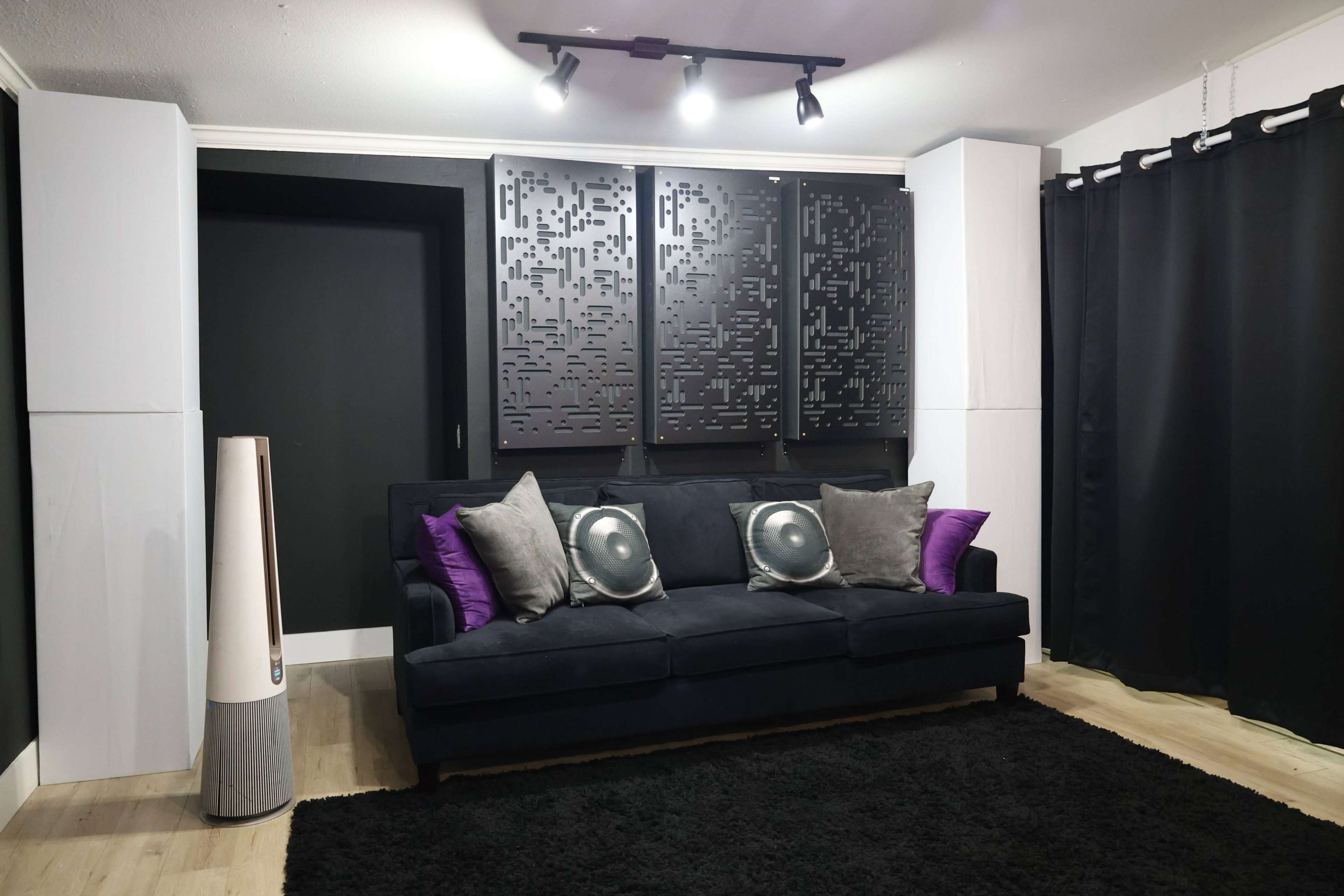 The image shows a modern living room with a black couch, decorative pillows, a dark rug, and a wall featuring stylish panels, along with curtains and a sleek floor lamp.
