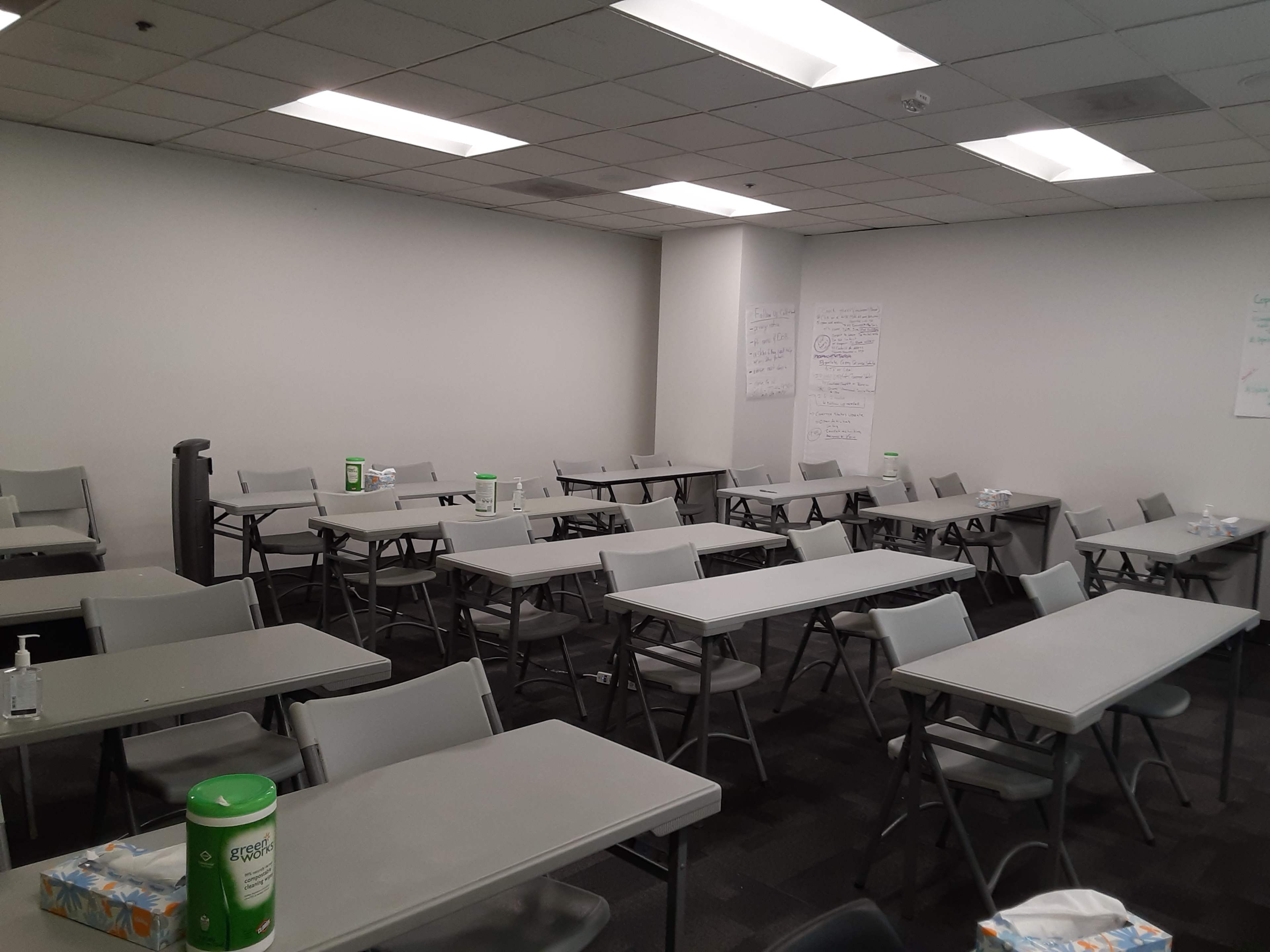 A classroom setting with multiple folding tables and chairs arranged in rows, along with some cleaning supplies and water bottles on the tables.