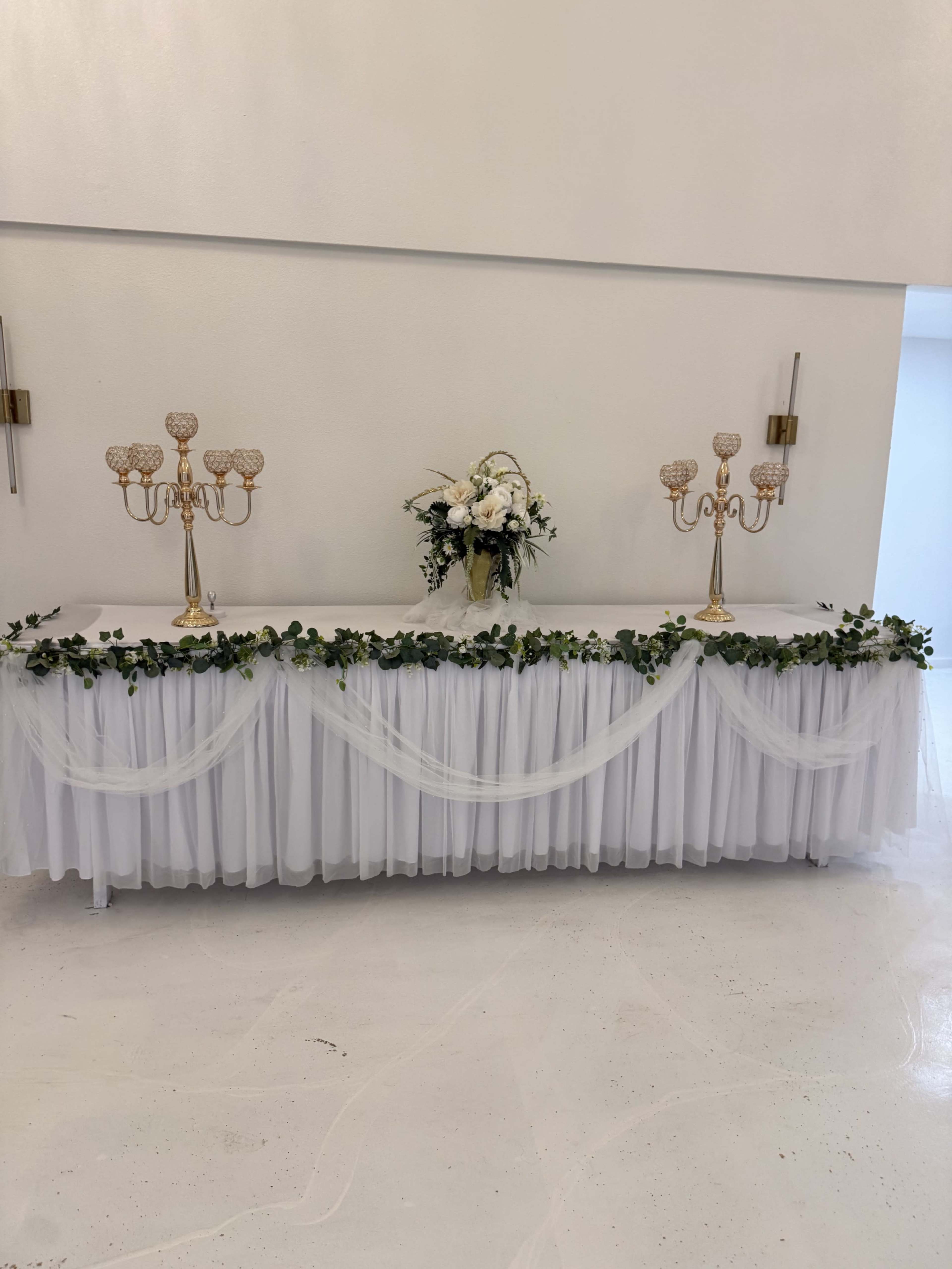 A long table is decorated with white draping, two gold candelabras, and a floral arrangement at the center.