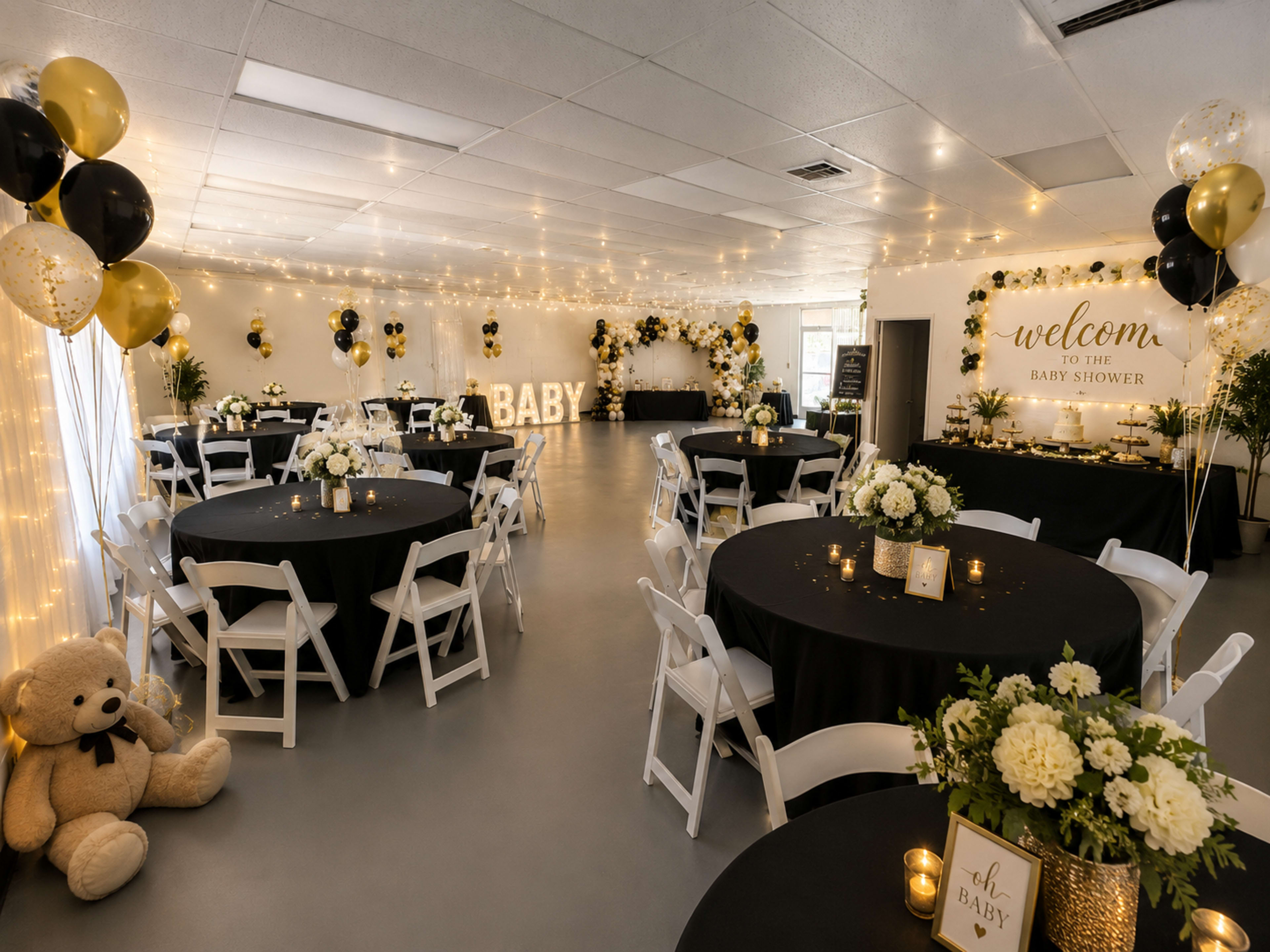 The room is decorated for a baby shower, featuring black and gold balloons, black tablecloths, and a teddy bear, with illuminated signs and floral arrangements on the tables.