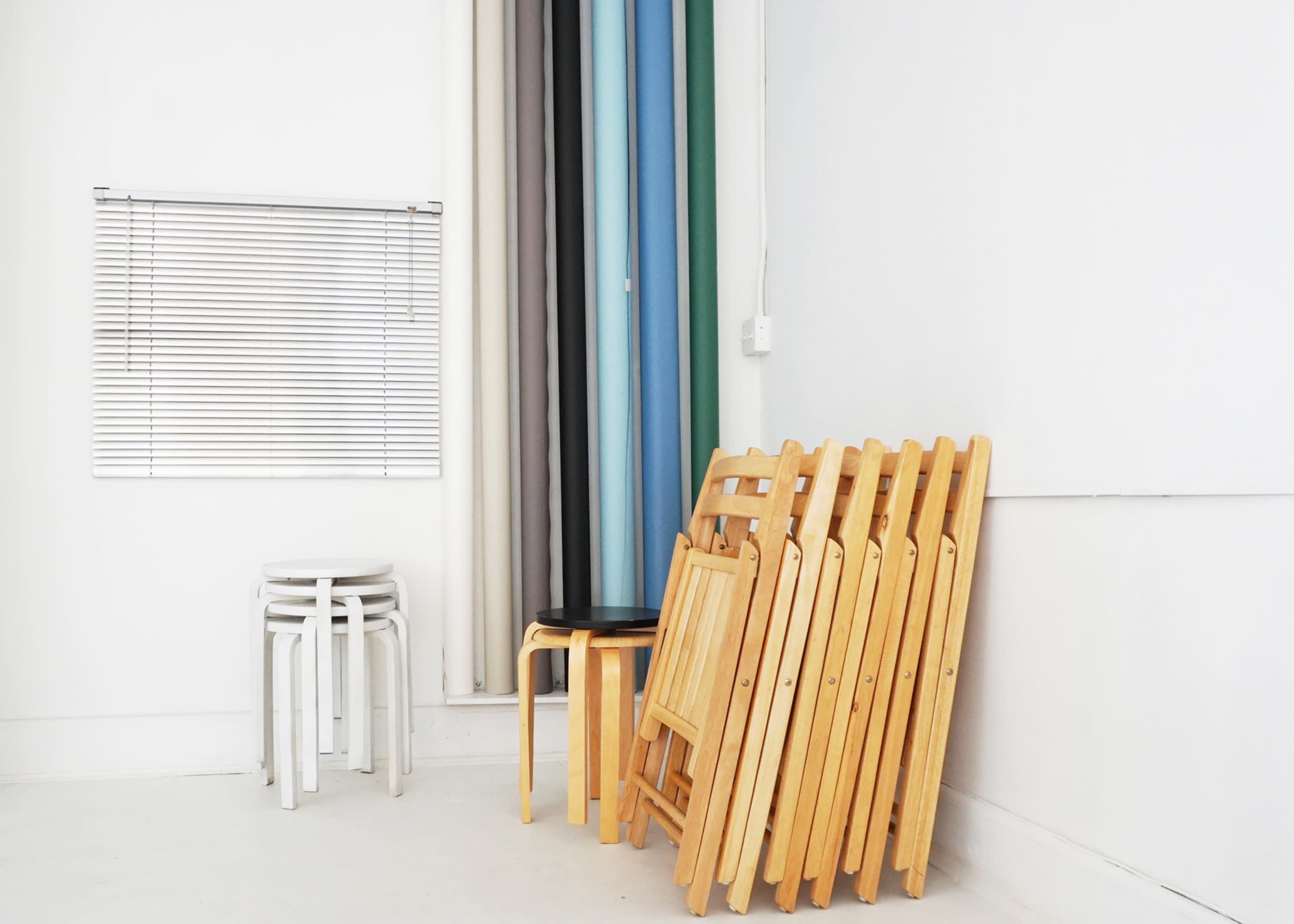 A stack of wooden chairs is positioned against a wall next to a window with blinds, alongside a white stool and colorful fabric rolls.