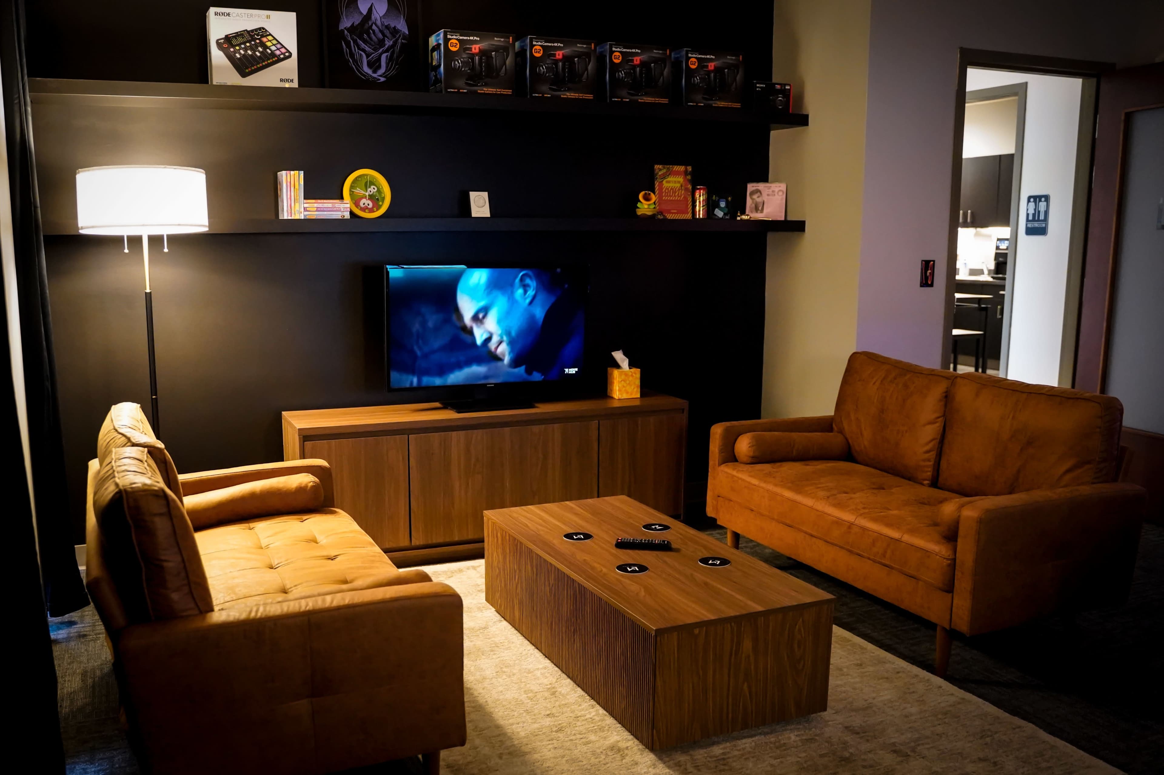 The scene features a cozy living area with two brown sofas facing a wooden coffee table, a television displaying a movie, and shelves showcasing various items and games.