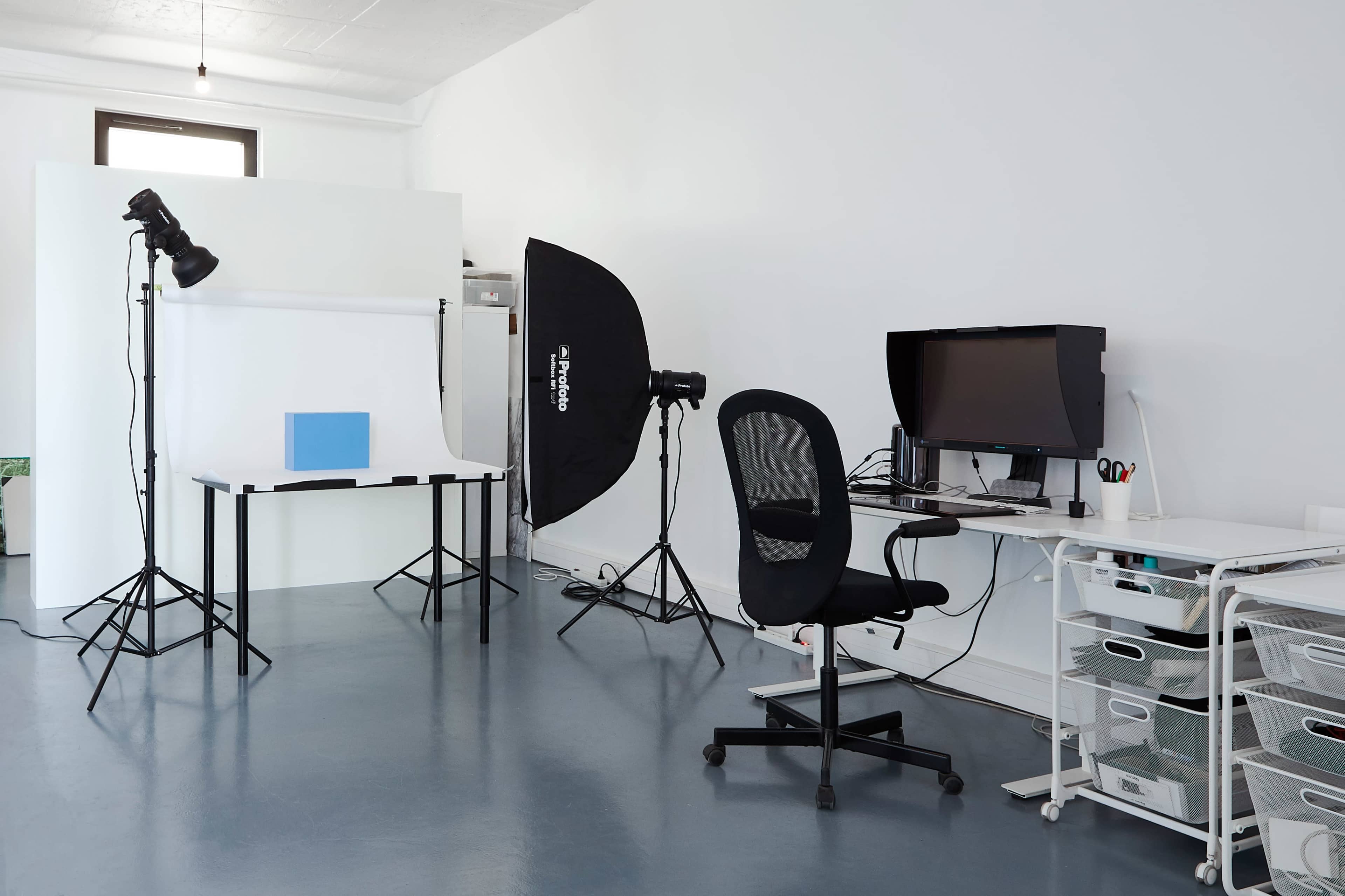 The image shows a minimalistic photography studio setup with a computer workstation, a backdrop, and various lighting equipment.
