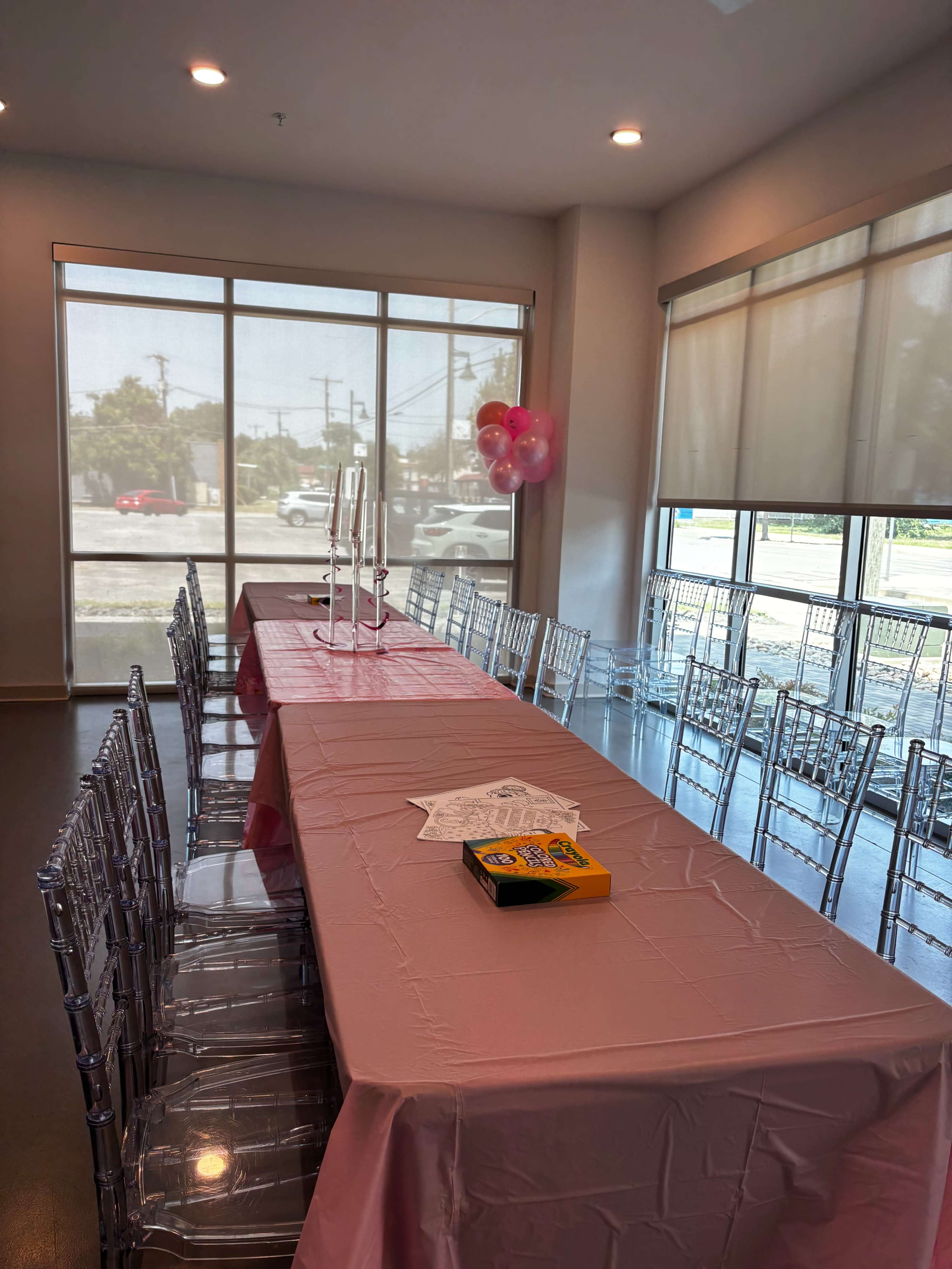 A long table covered with a pink tablecloth is set in a brightly lit room, surrounded by clear acrylic chairs and decorated with balloons in the corner.