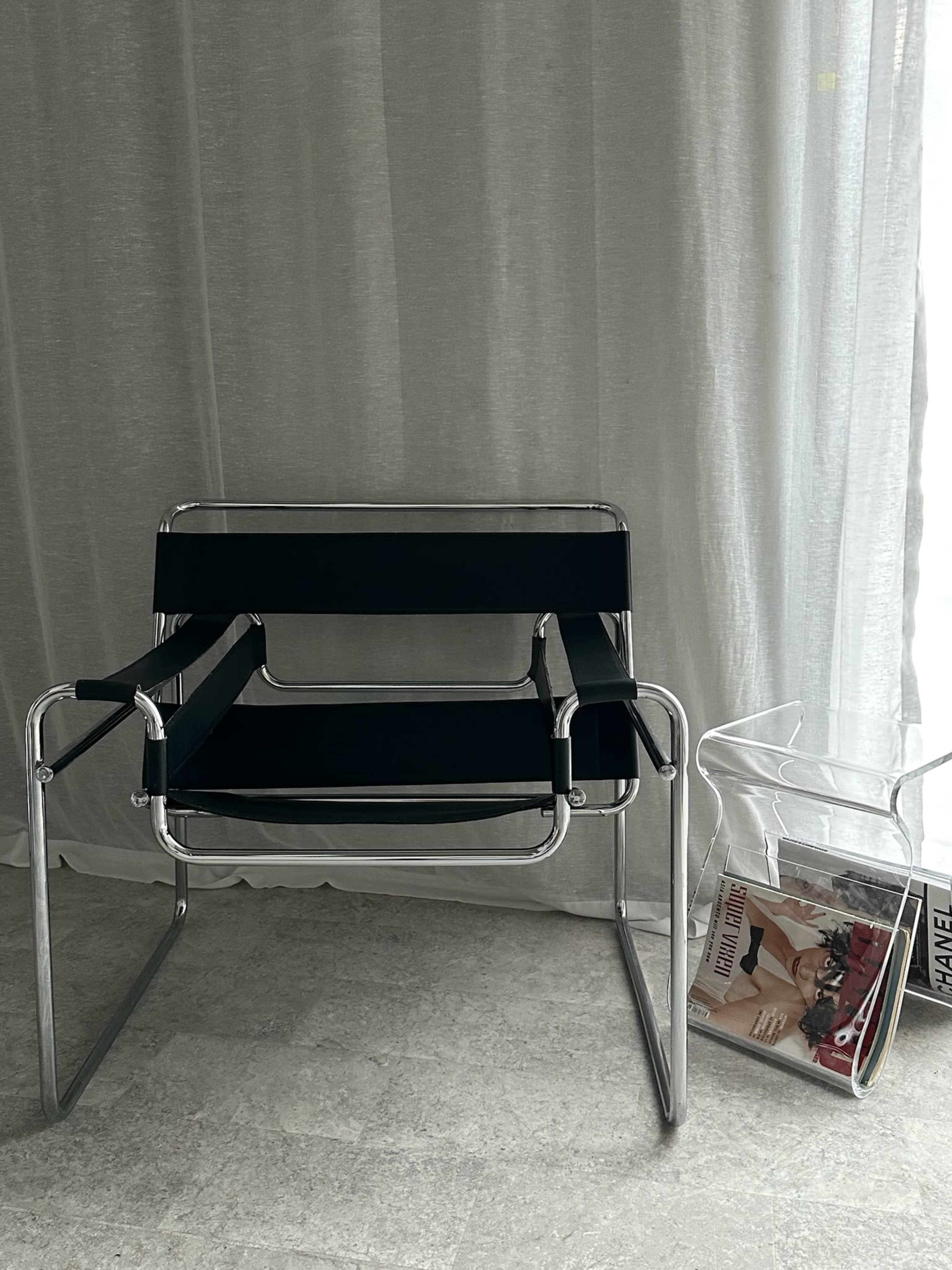 A modern black chair with chrome frames sits beside a clear acrylic side table holding magazines, in front of a flowing white curtain.