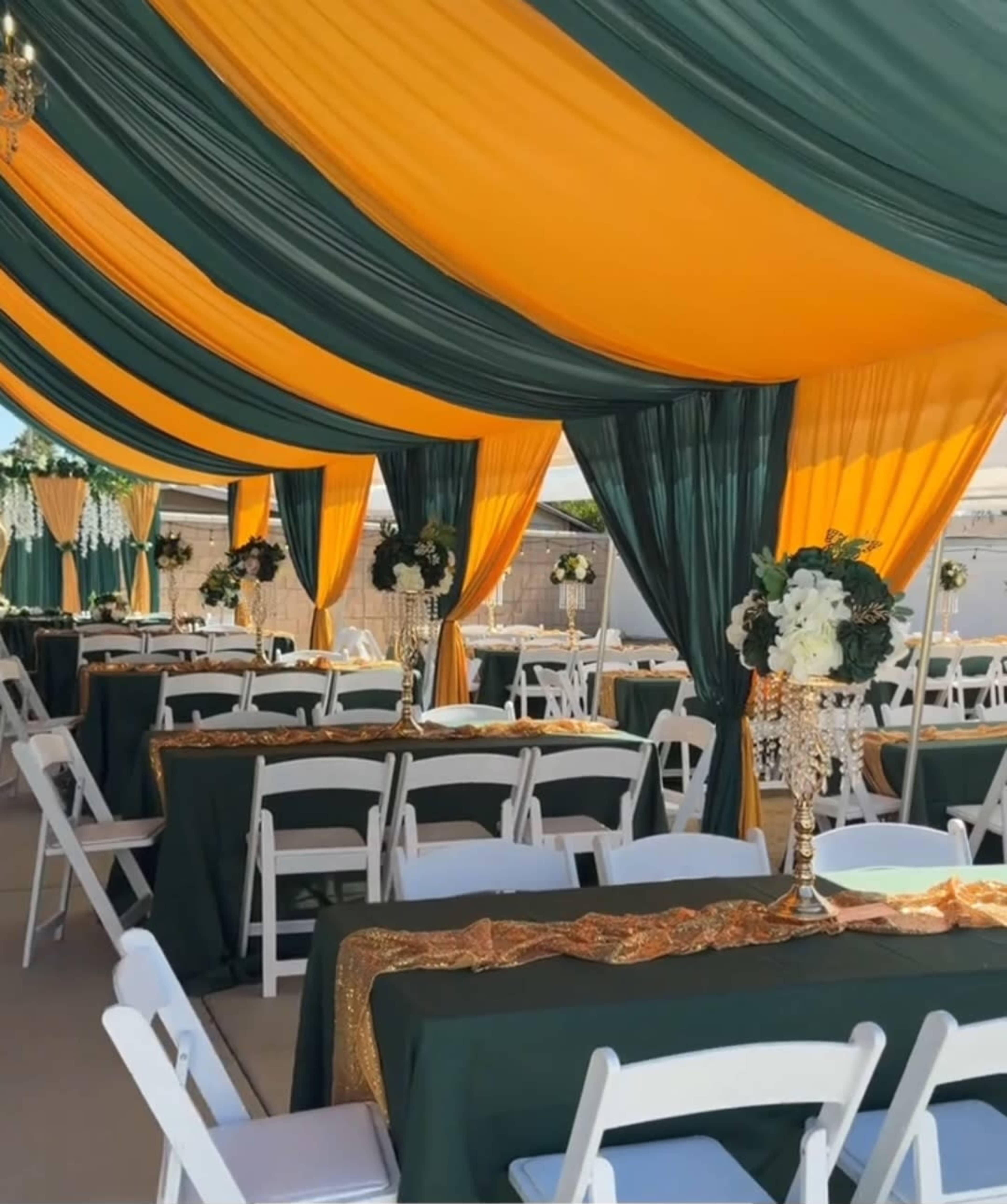 The image shows a decorated tent with green and yellow draped fabric overhead and tables set with white chairs and tablecloths adorned with gold accents.