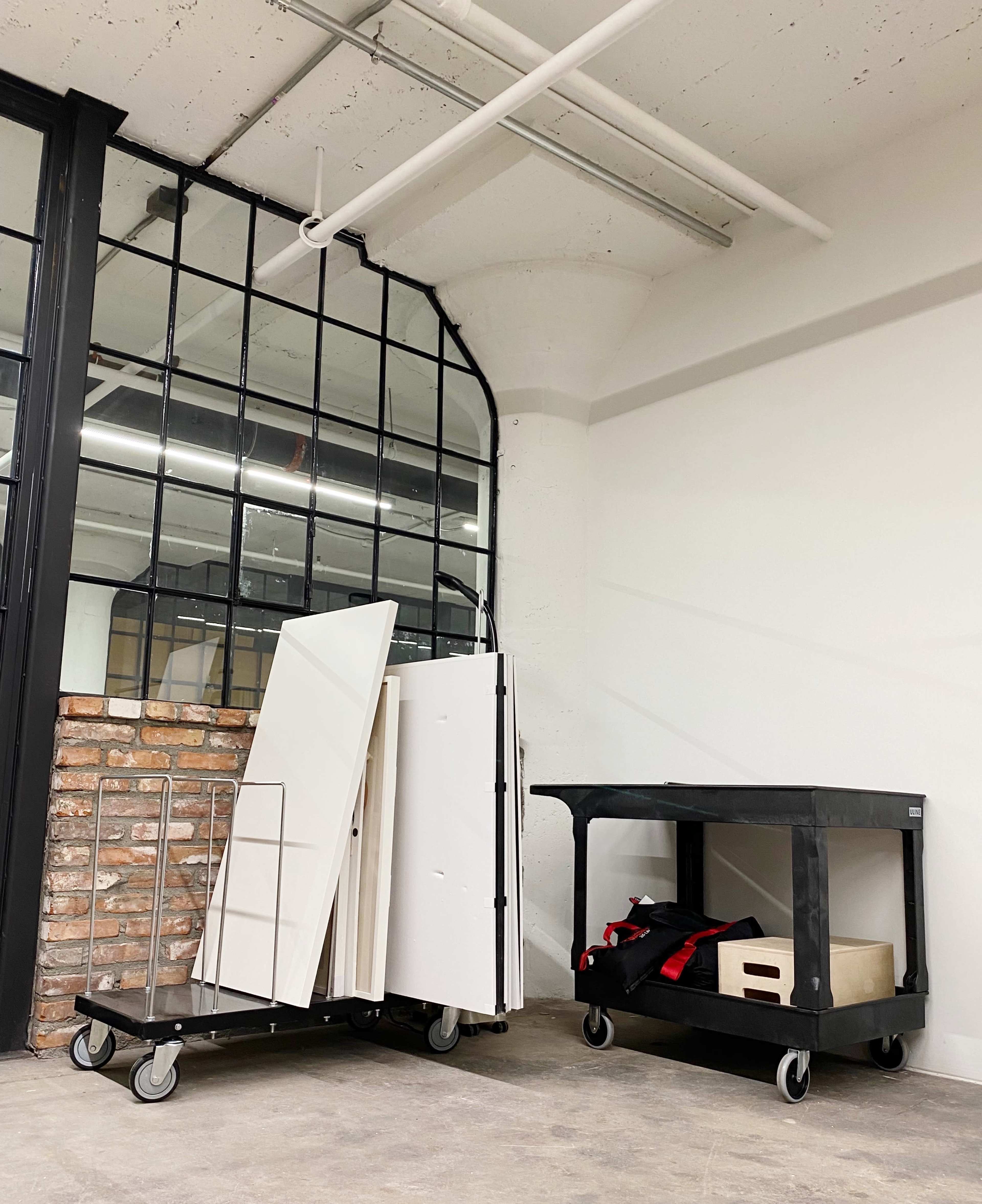 A black cart and a wheeled trolley with stacked panels and boxes are positioned against a brick wall in an industrial space.