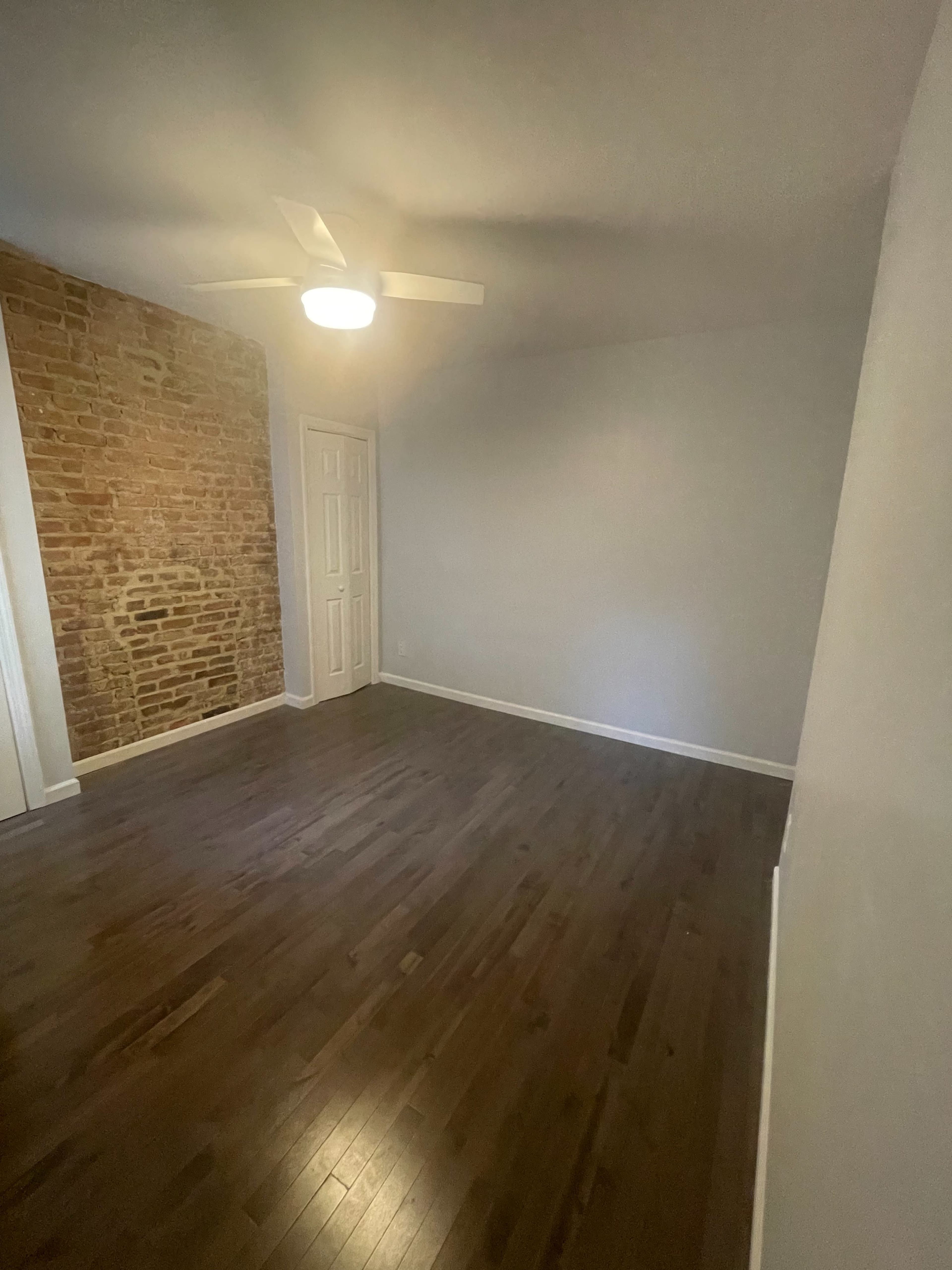 A spacious room with a wooden floor, a ceiling fan, and a brick accent wall.