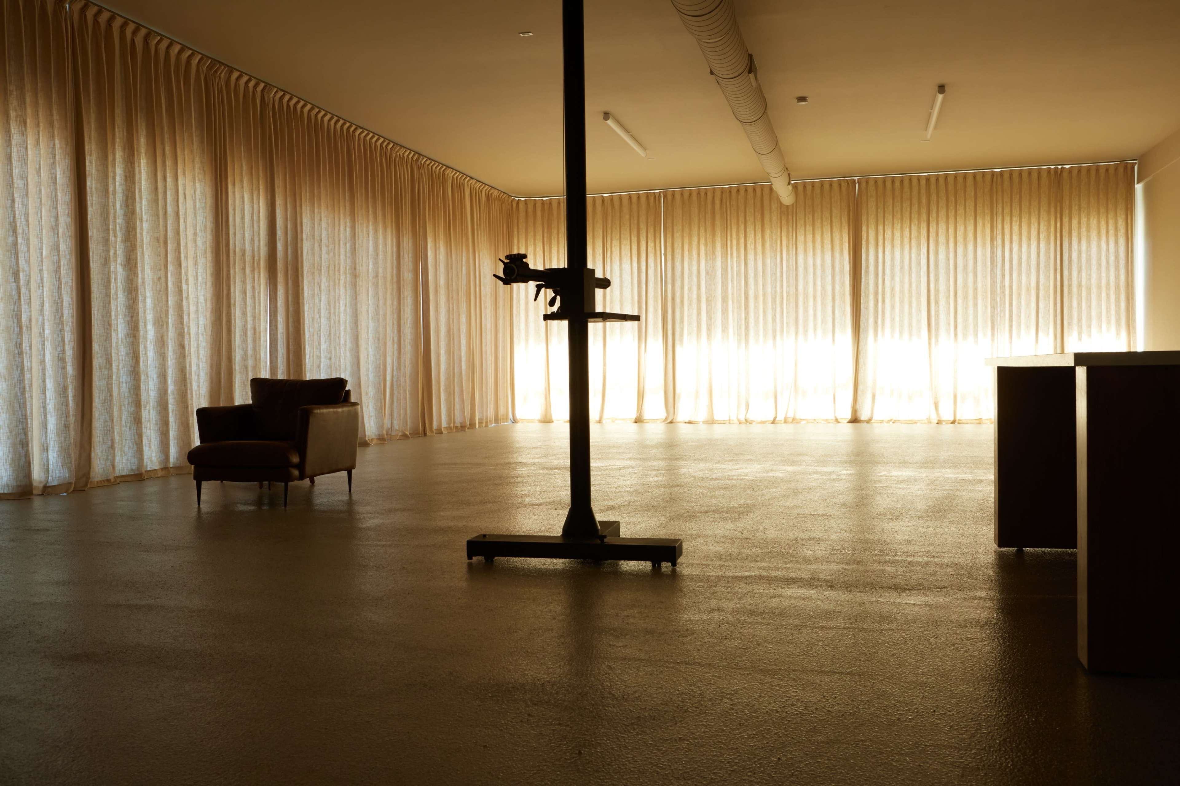 A spacious room features large windows with sheer curtains, a brown armchair, and a tall, industrial-style object in the center.
