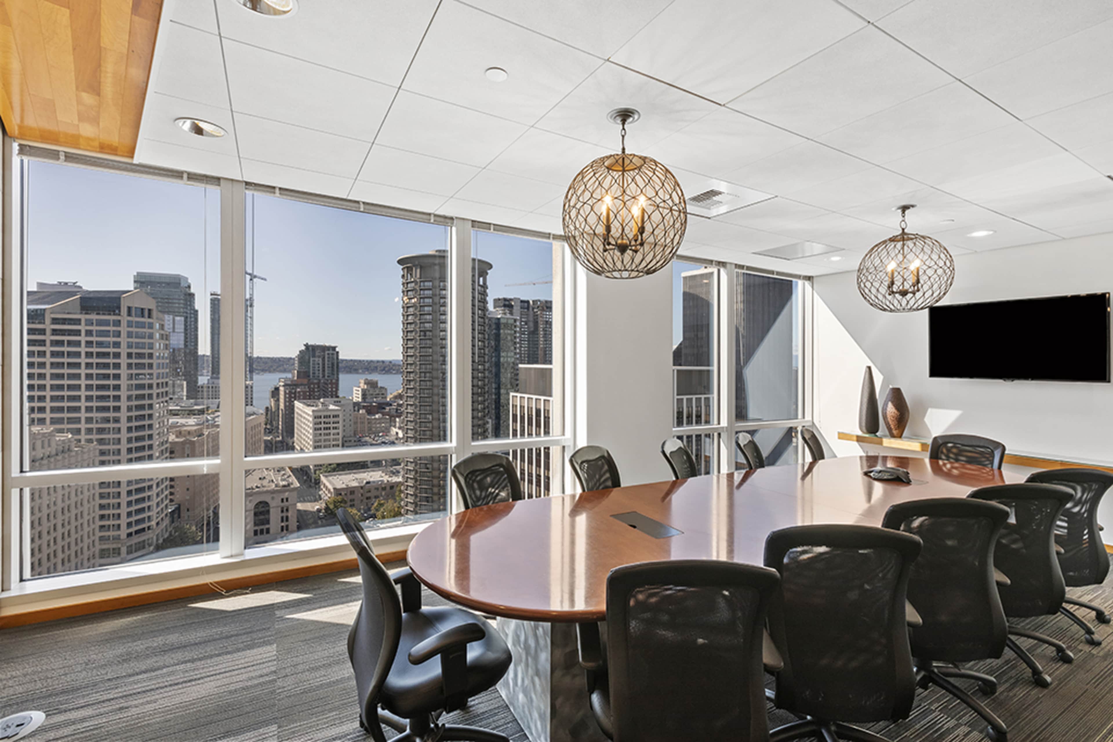 A modern conference room features a large wooden table surrounded by black chairs, with floor-to-ceiling windows showcasing a city skyline and waterfront view.
