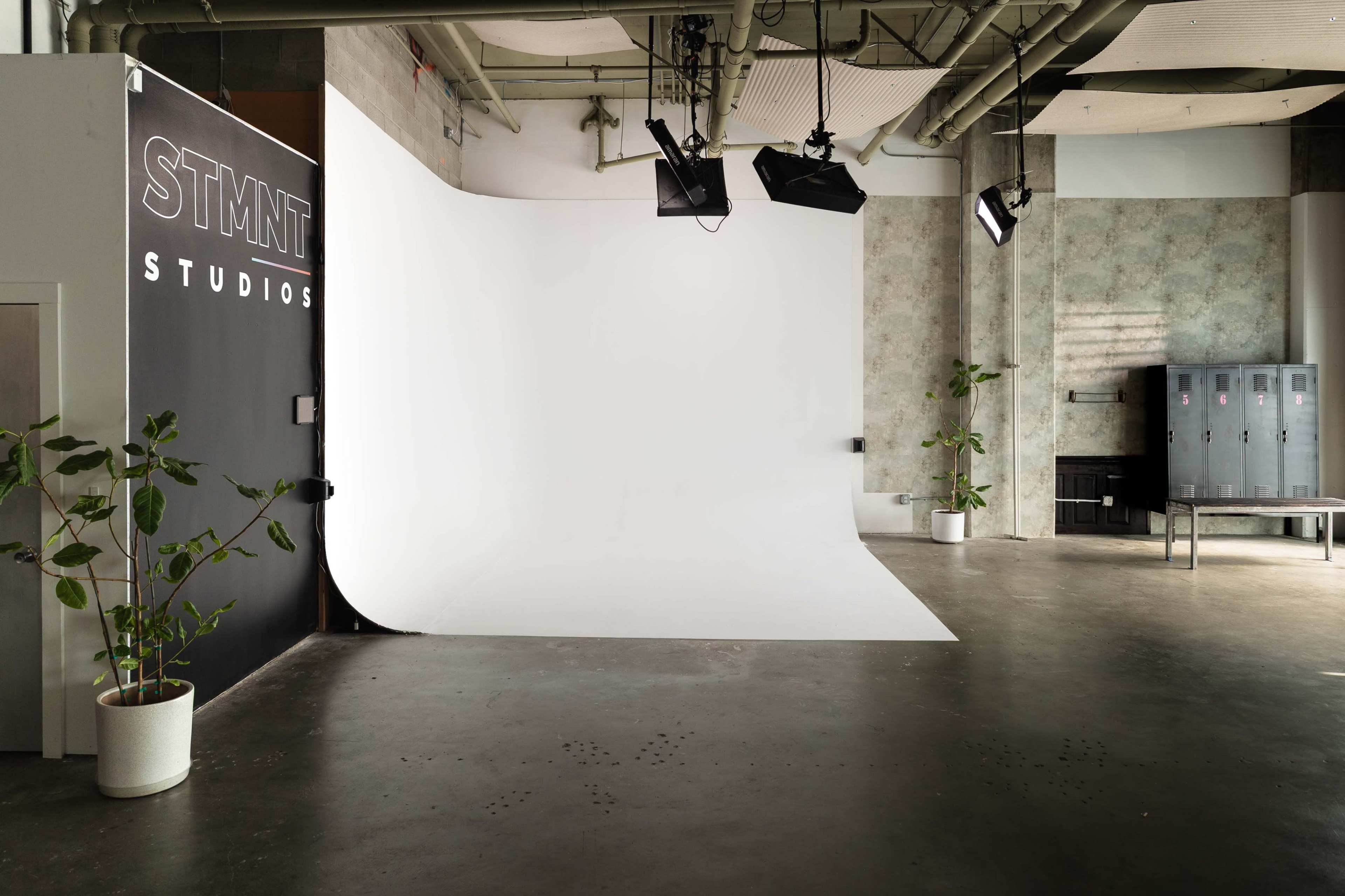 The image shows a spacious studio with a white backdrop, modern lighting, a black wall featuring the text "STMT STUDIOS," and potted plants.