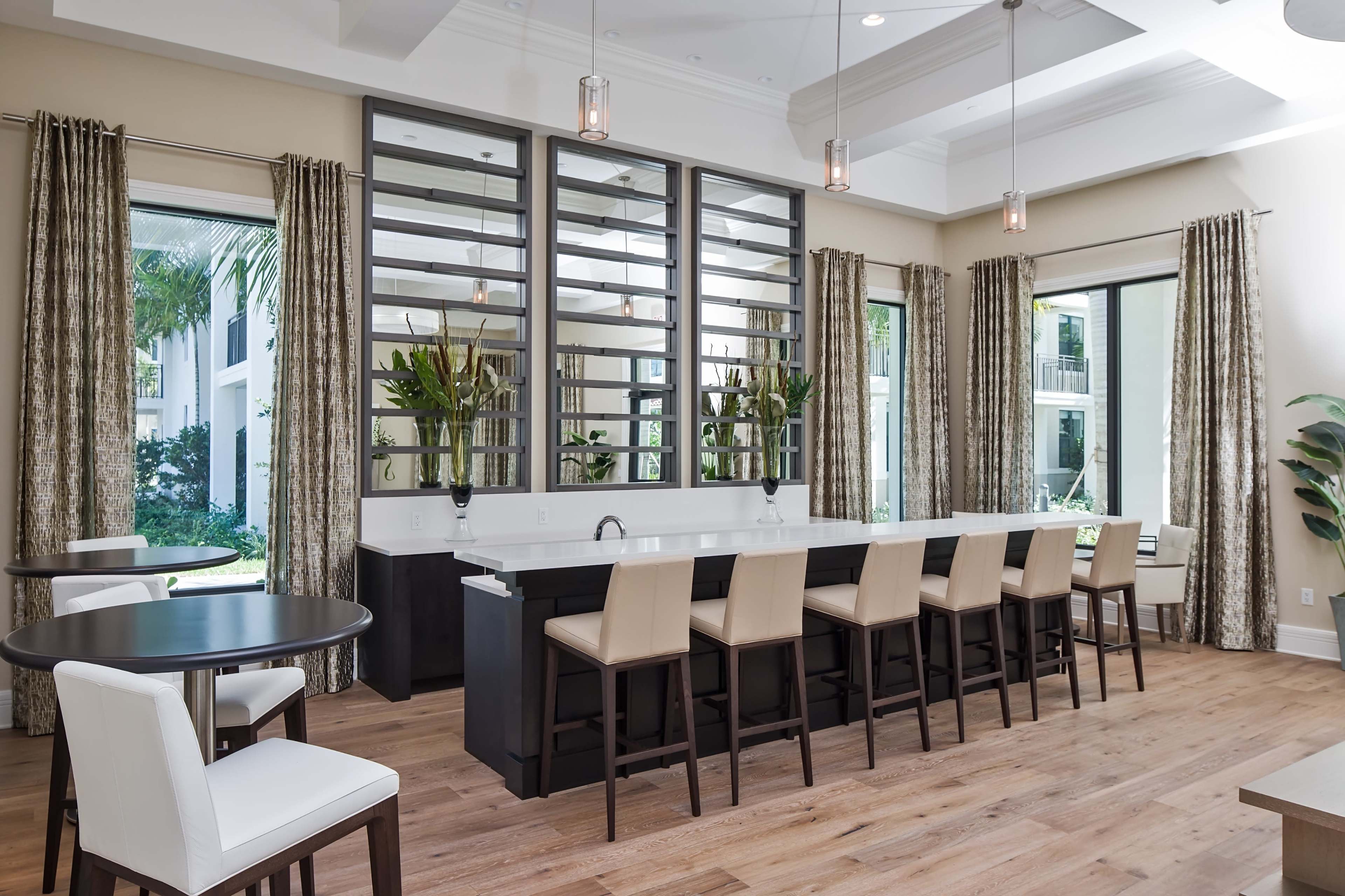 A modern kitchen area features a long bar with beige stools, surrounded by large windows and lush greenery visible outside.