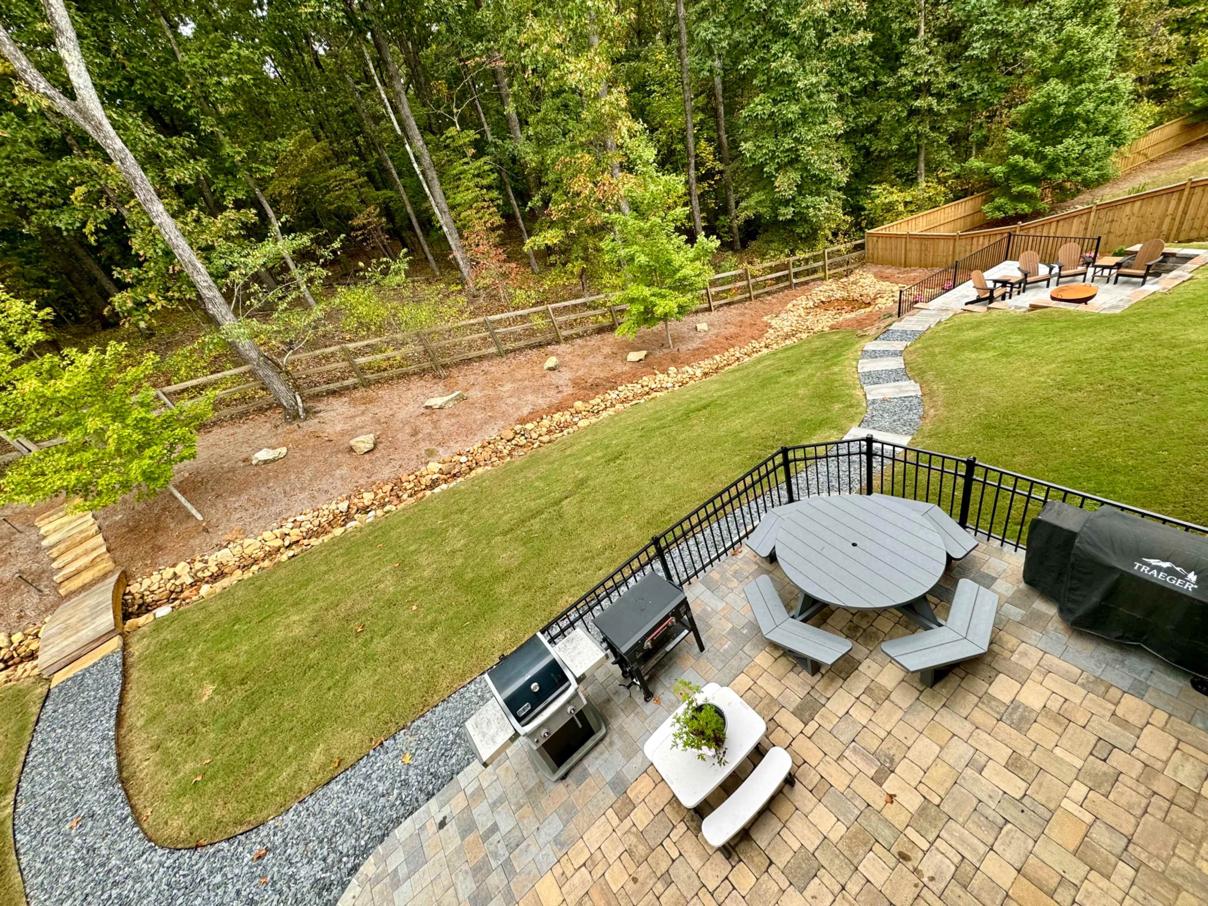 An outdoor patio features a circular table and chairs, a grill, and a pathway leading to a lawn bordered by trees and a wooden fence.