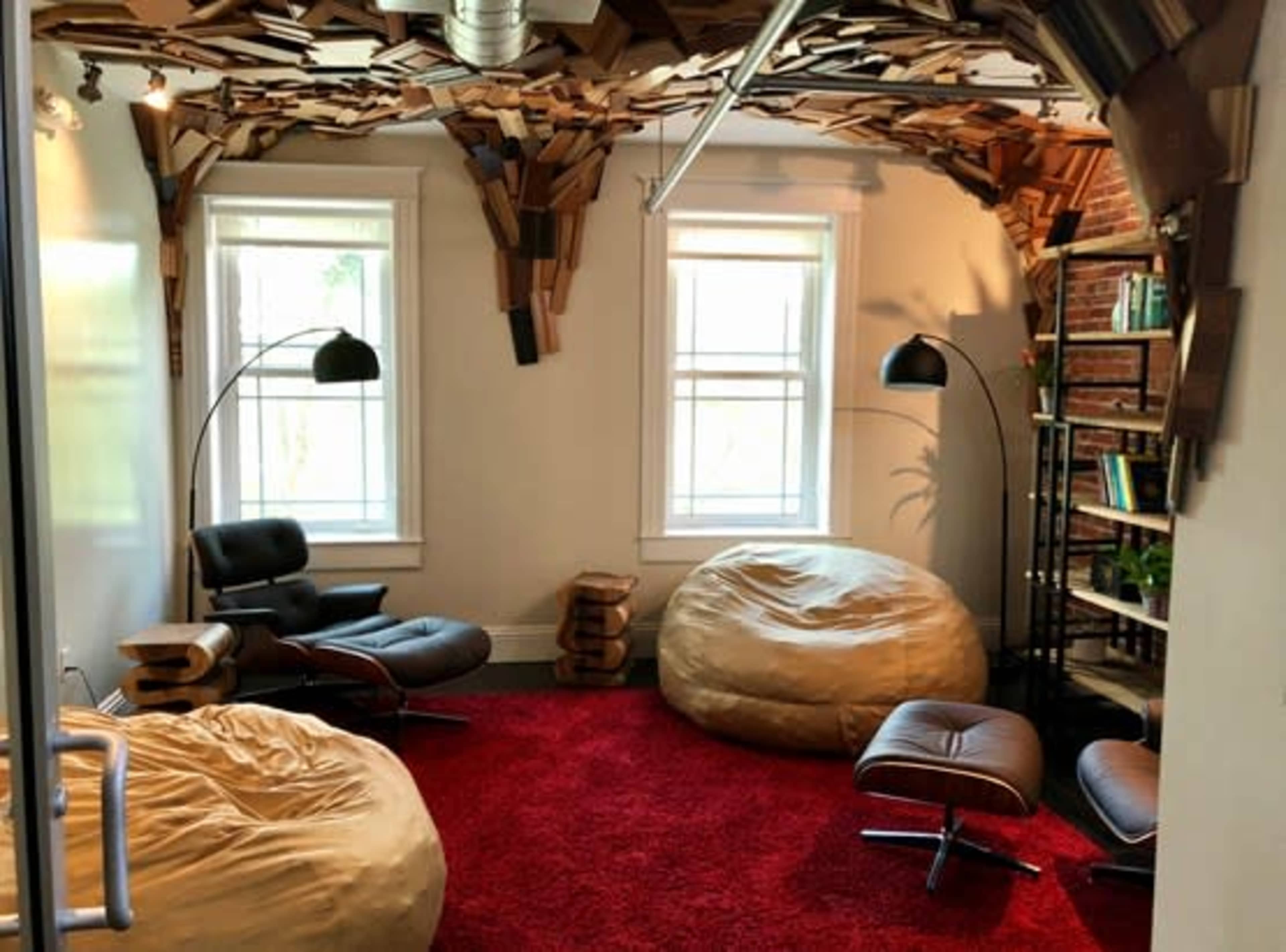 The room features a red carpet, two large bean bags, a black lounge chair, and a wooden bookshelf against a wall with a ceiling made of stacked cardboard pieces.