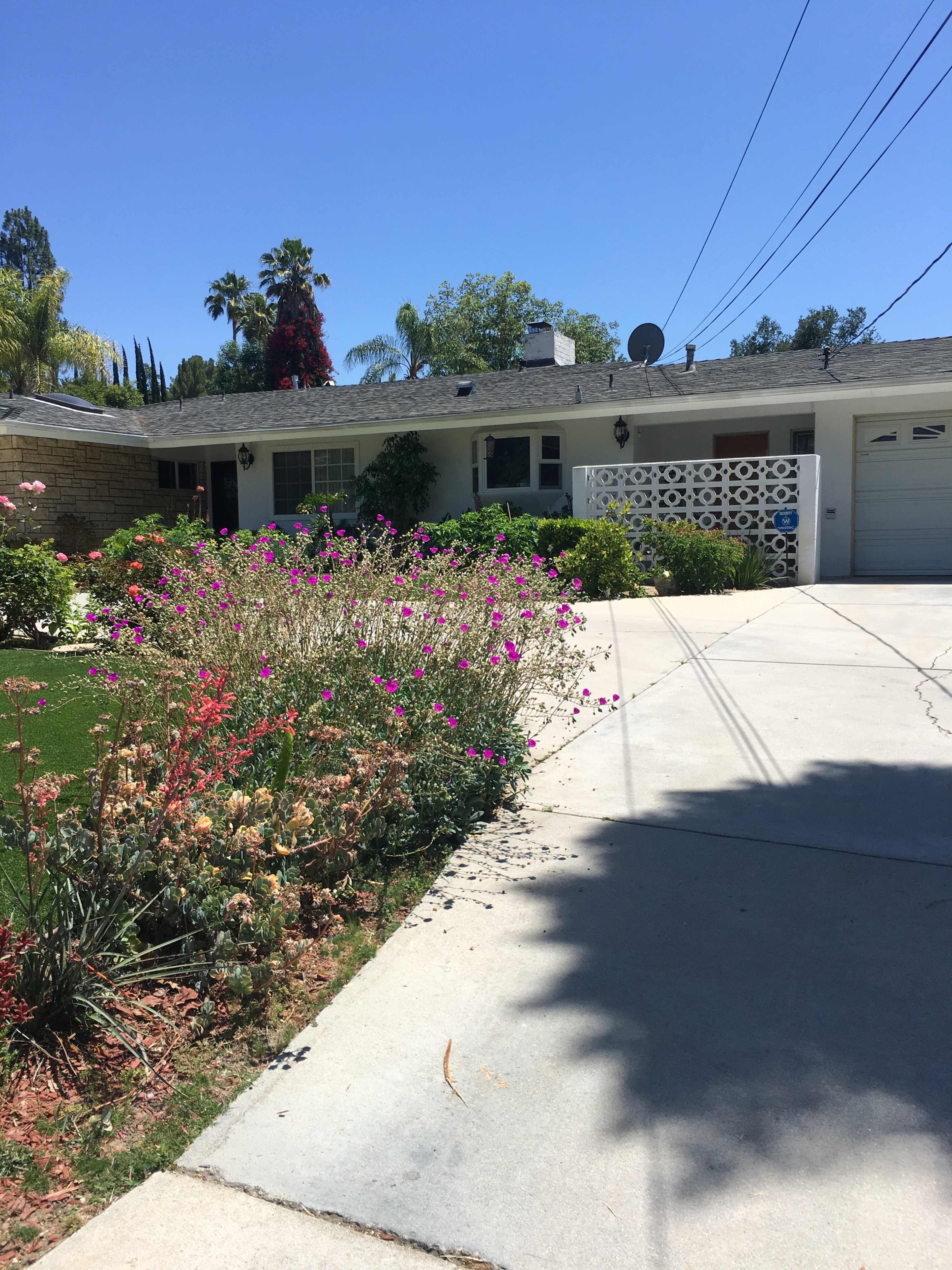 The image shows a residential house with a landscaped front yard featuring colorful flowers and a paved driveway.
