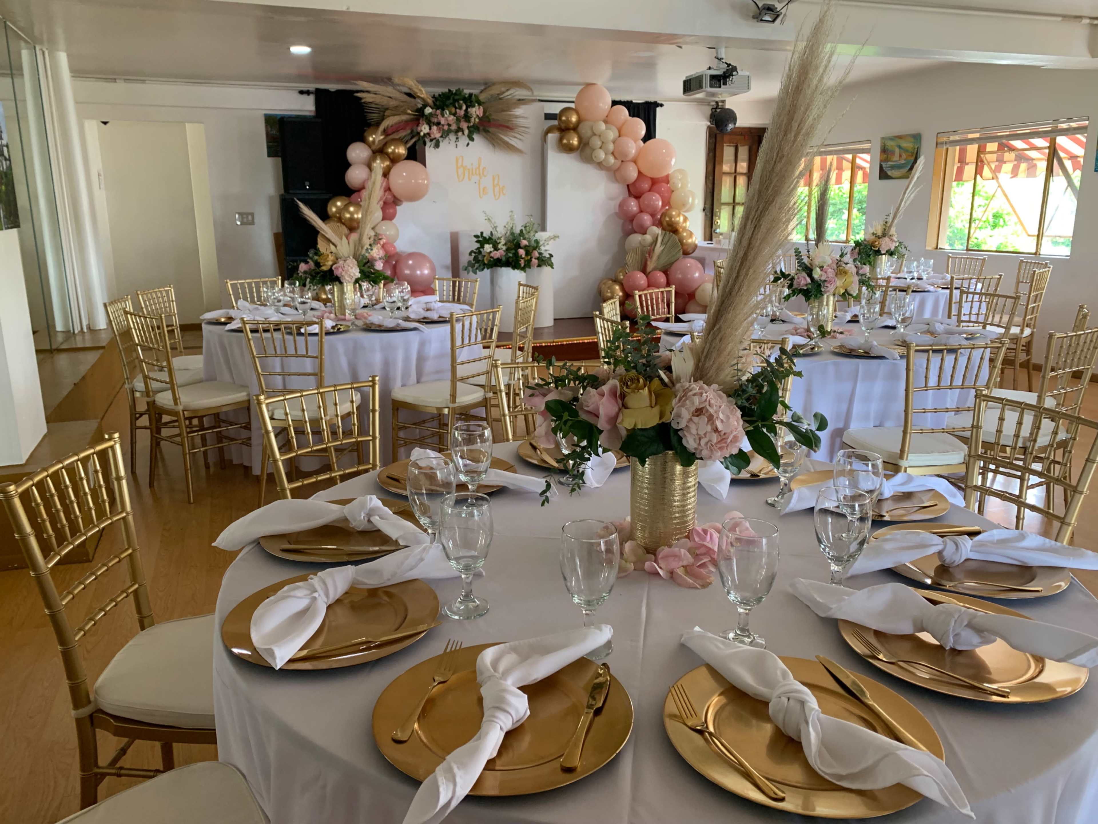 The image shows an elegantly arranged event space with gold-accented tables and floral decorations, ready for a celebration.