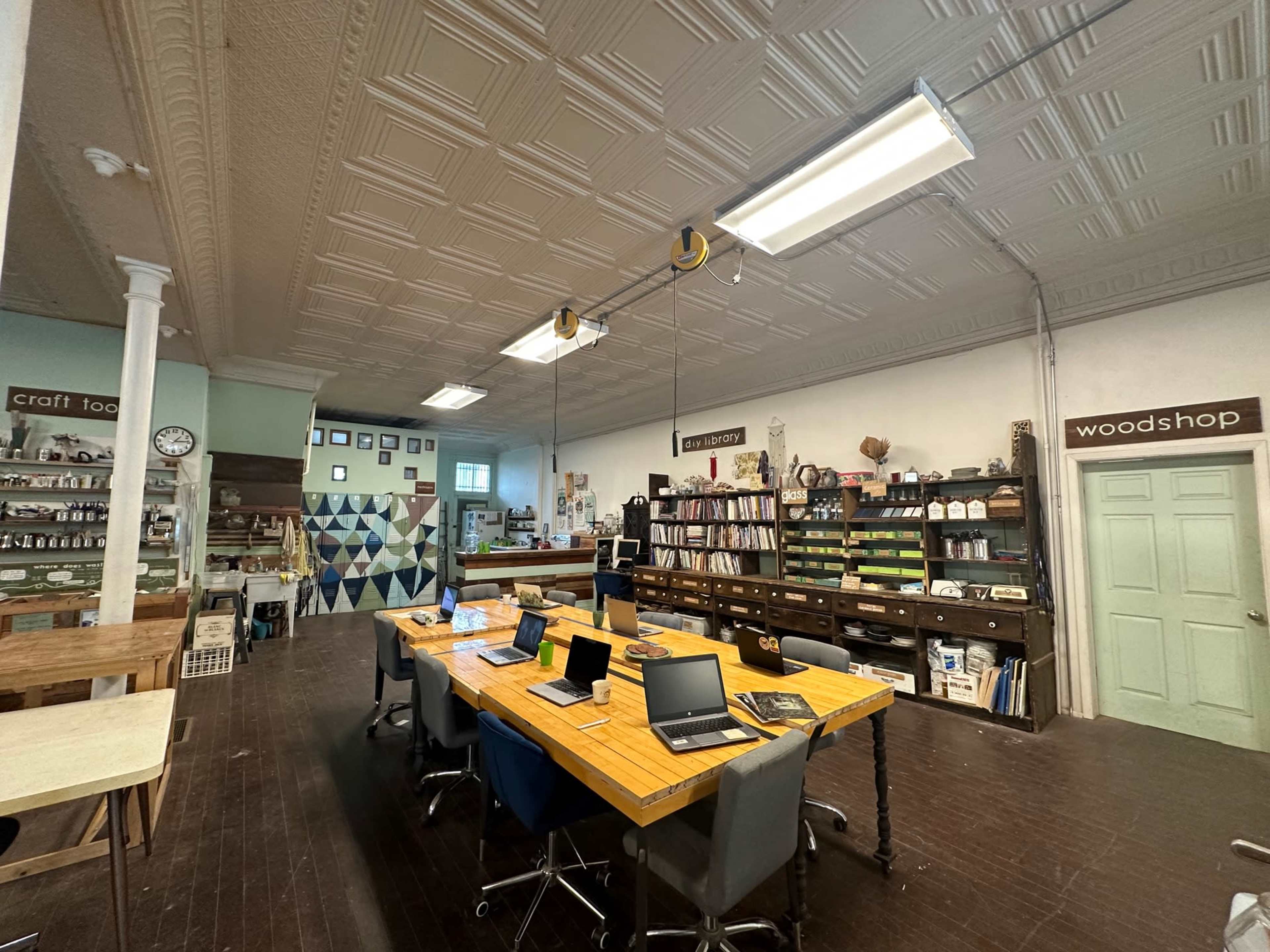 A spacious workshop with a long wooden table surrounded by laptops, bookshelves filled with materials, and various crafting supplies along the walls.