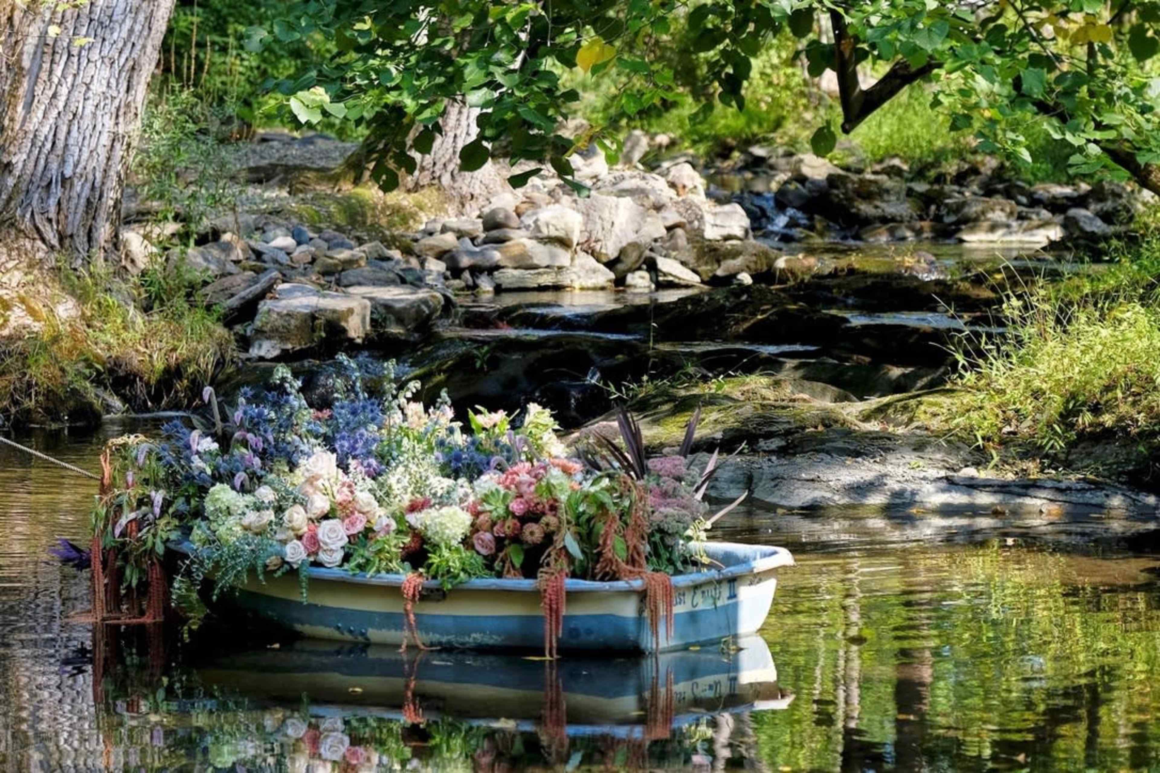 A small boat filled with various flowers floats on a calm stream surrounded by greenery and rocks.