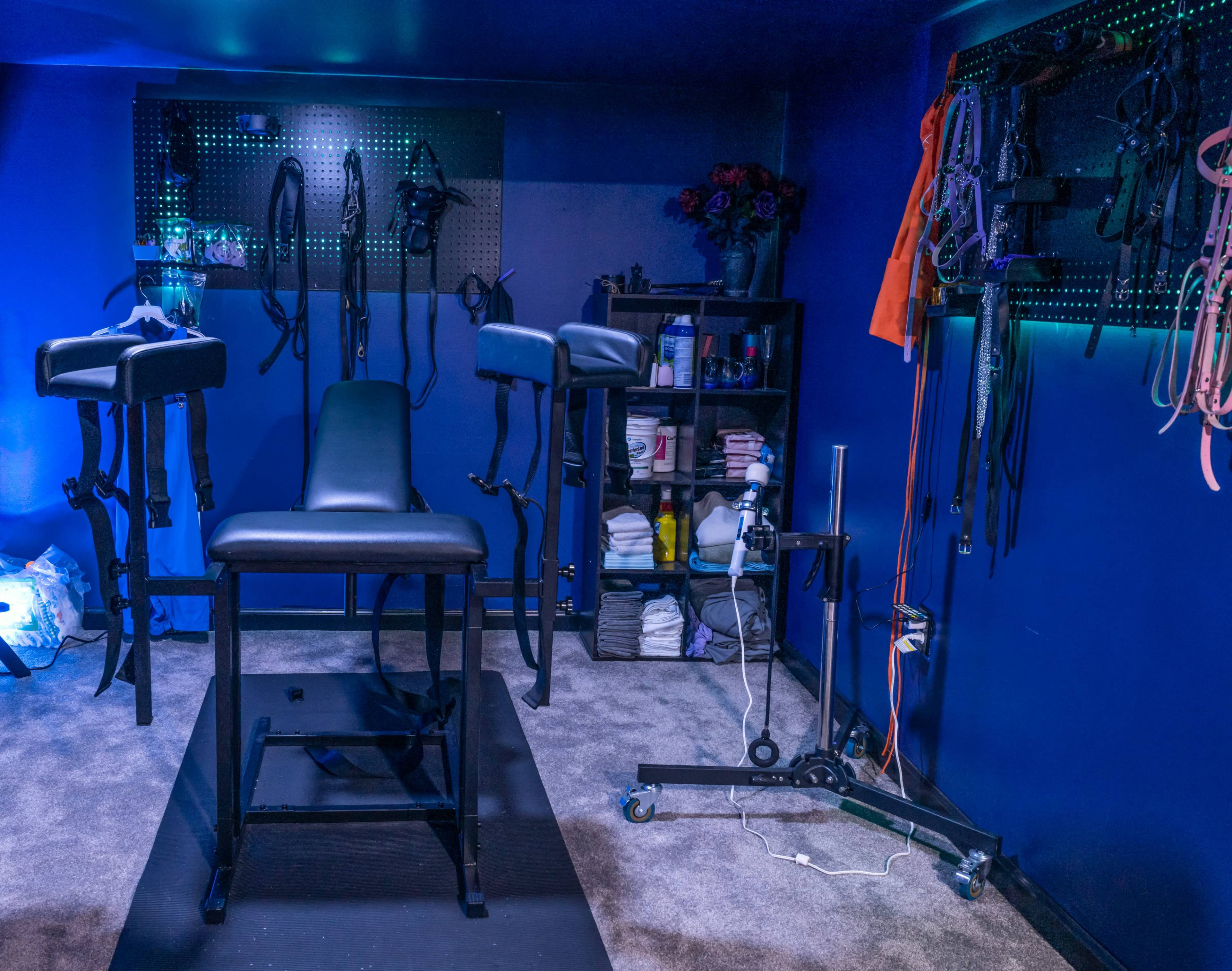 The image depicts a room with blue walls containing various pieces of equipment, including a central black chair and straps mounted on the walls.