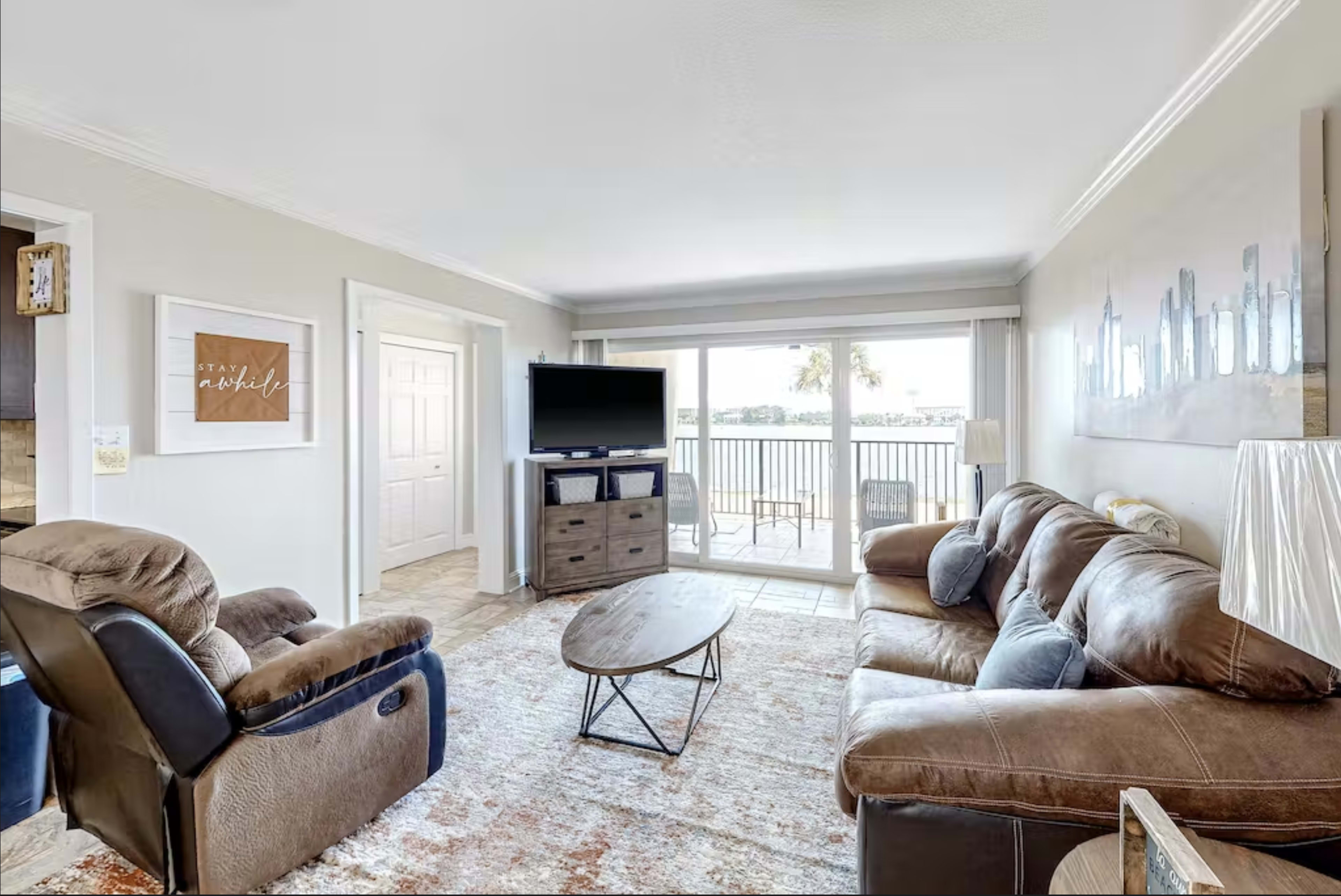 The living room features a brown leather sofa and recliner, a coffee table, a television, and large windows with a view of the outdoors.