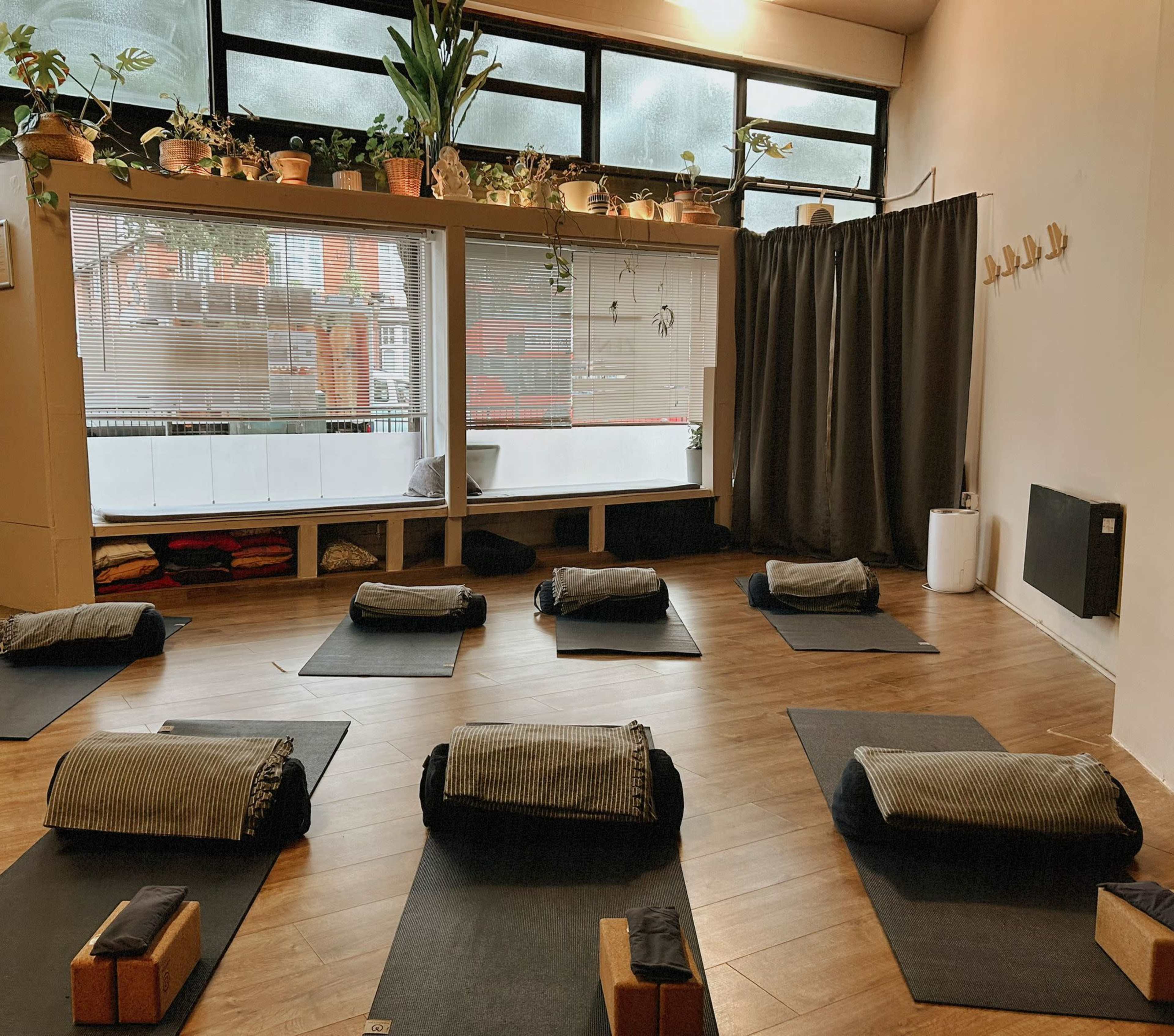 The image shows a serene yoga studio with mats, bolsters, and blocks neatly arranged on a wooden floor, surrounded by large windows with plants and a curtain in the background.