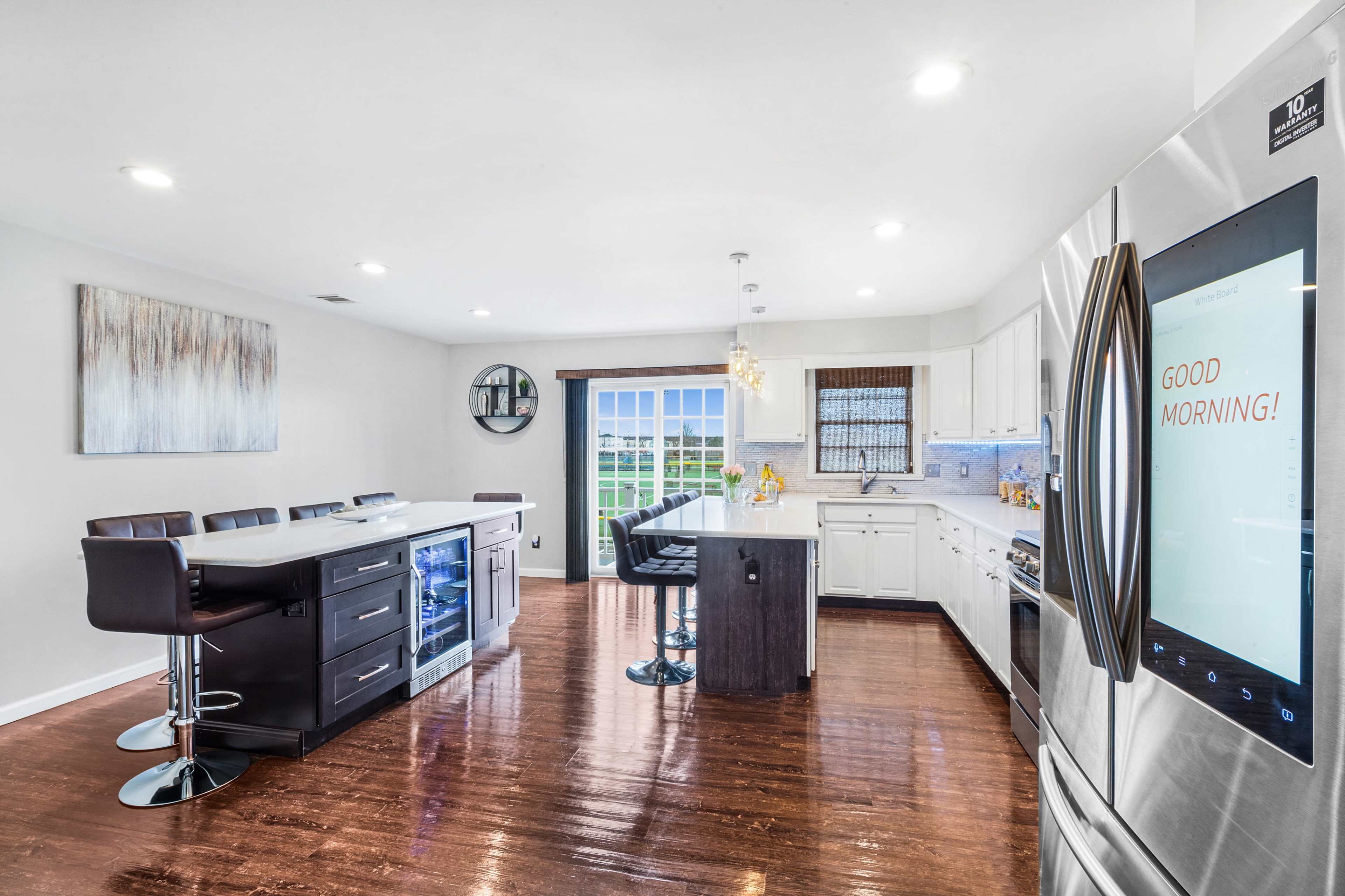 A modern kitchen features white cabinets, an island with bar seating, a stainless steel refrigerator, and large windows overlooking an outdoor view.