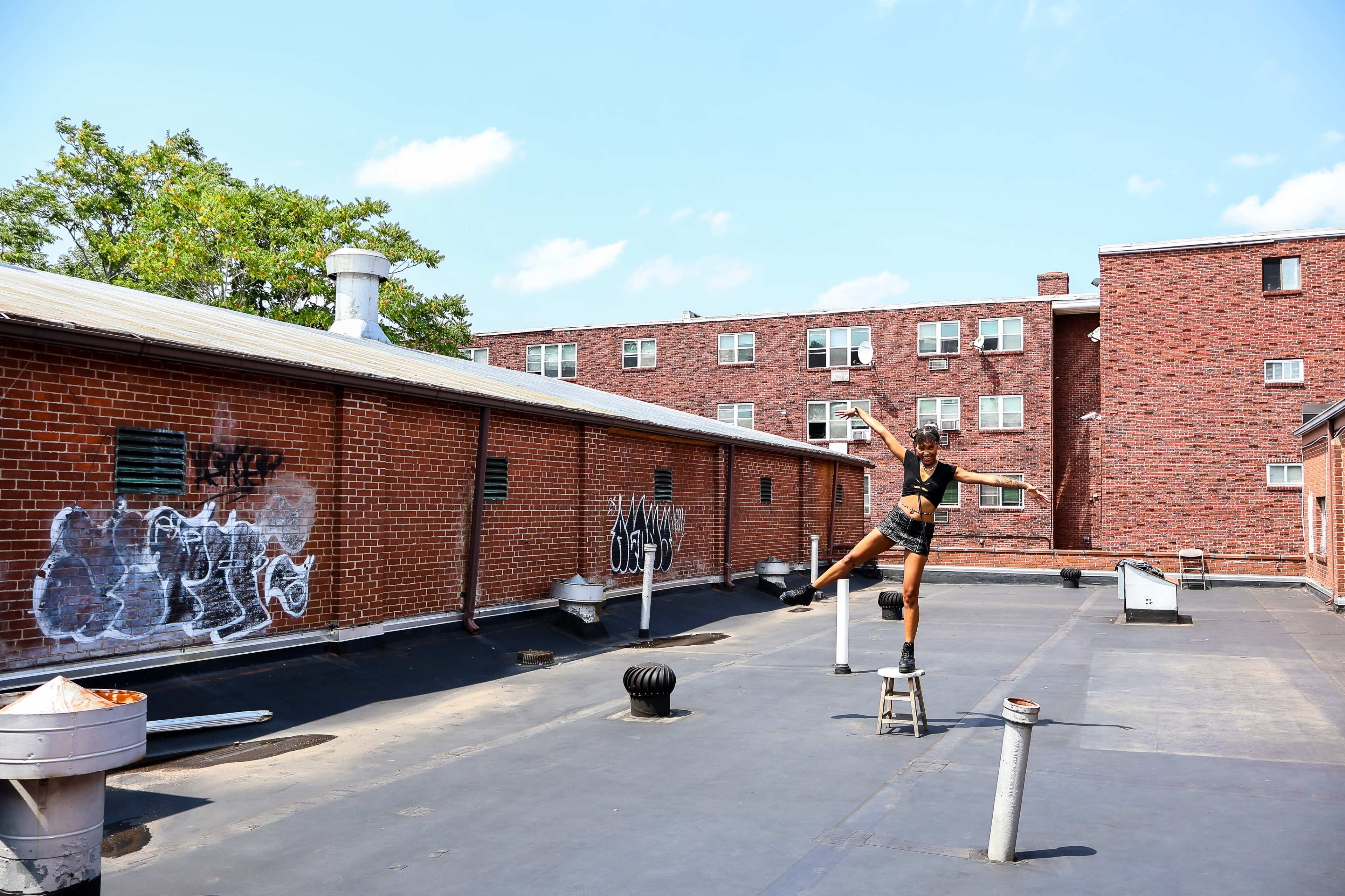 A person balances on a stool while performing a trick on a rooftop surrounded by brick buildings.