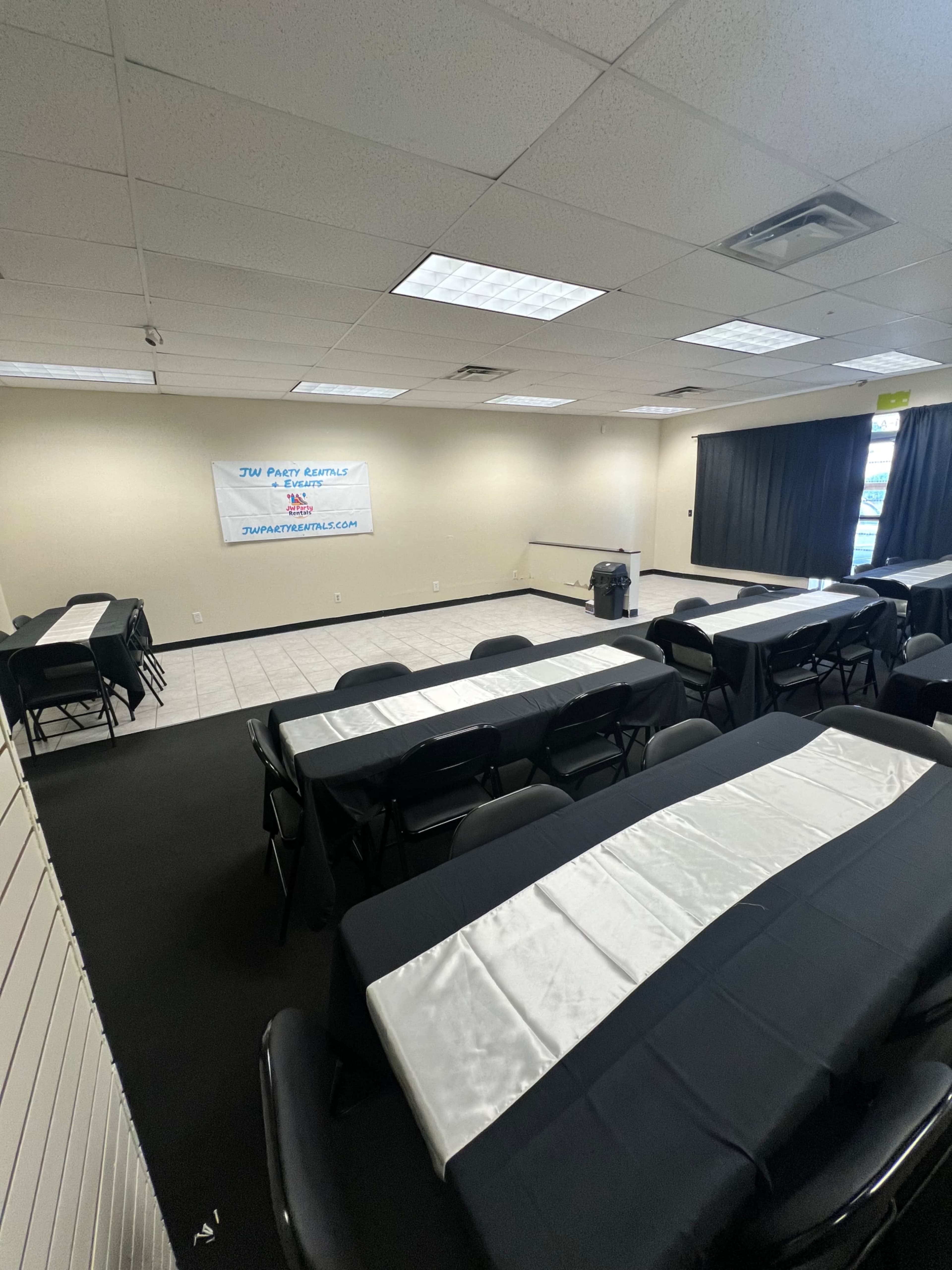 The image shows an empty indoor space set up with rows of tables covered in black and white tablecloths, with a banner displaying "JMU Party Rentals" on one wall.