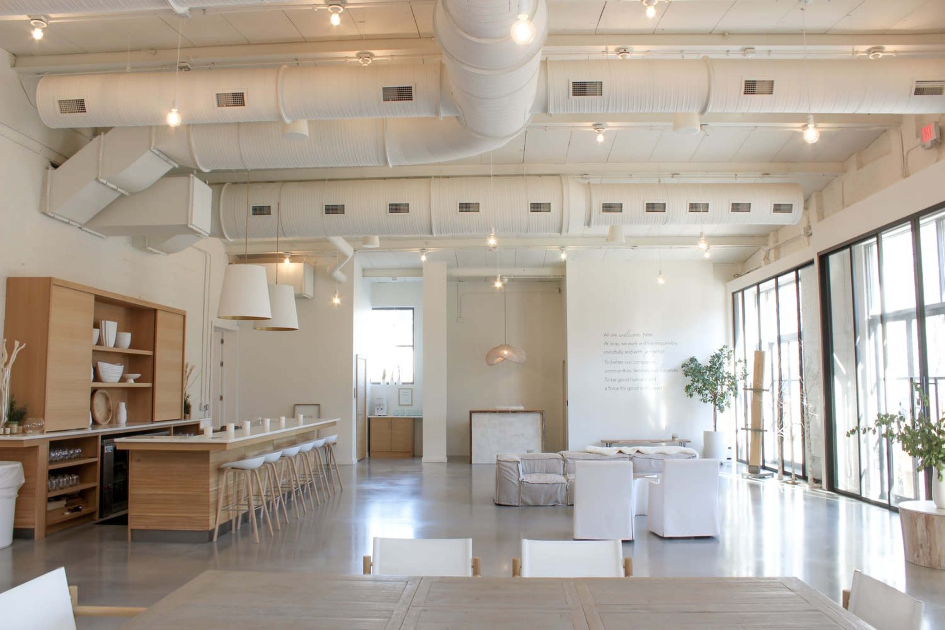 A spacious, modern interior with high ceilings, large windows, and minimalist furniture arranged in a dining and lounging area.