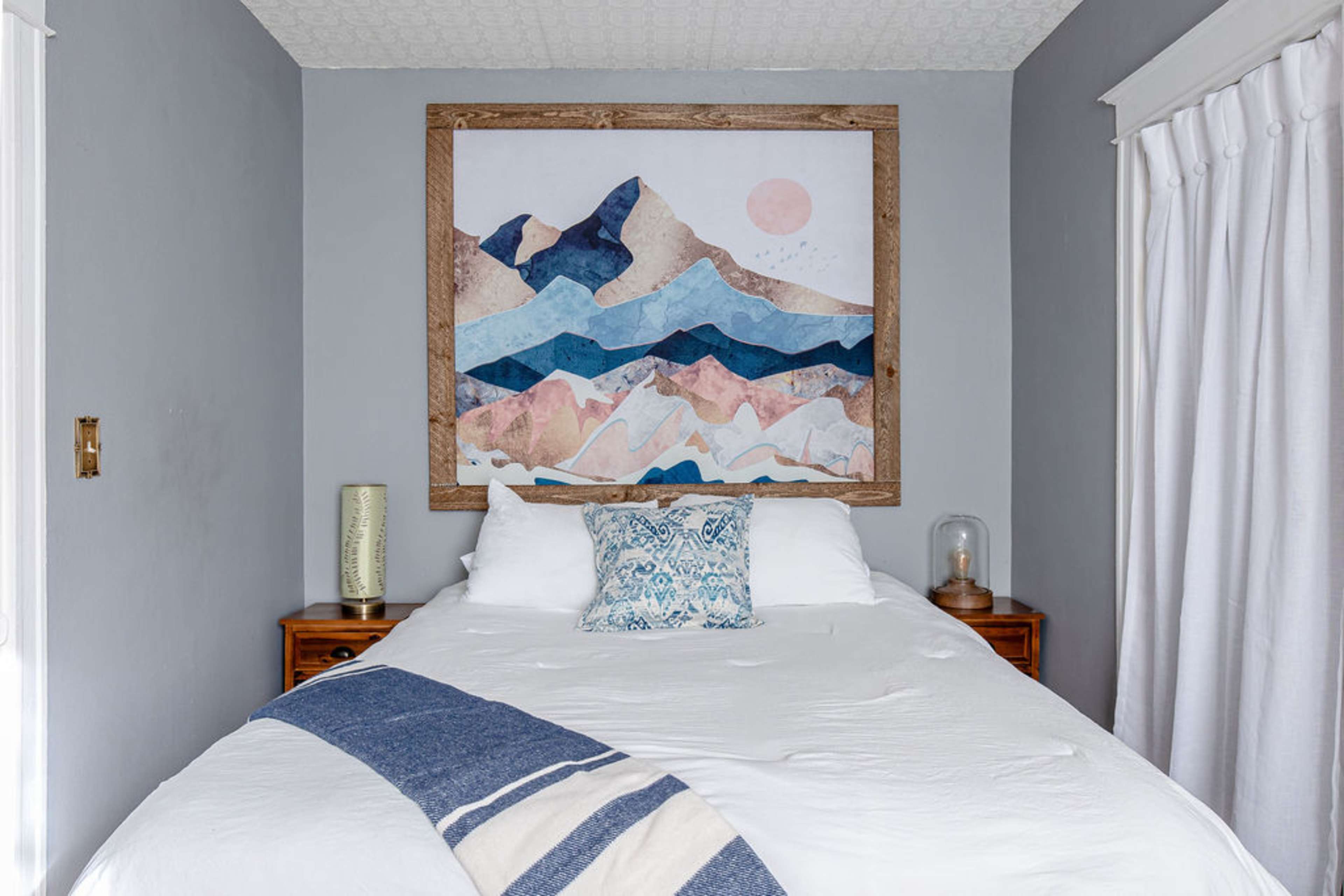 A bedroom features a bed with a white comforter, a decorative pillow, and a large mountain-themed artwork above the headboard.