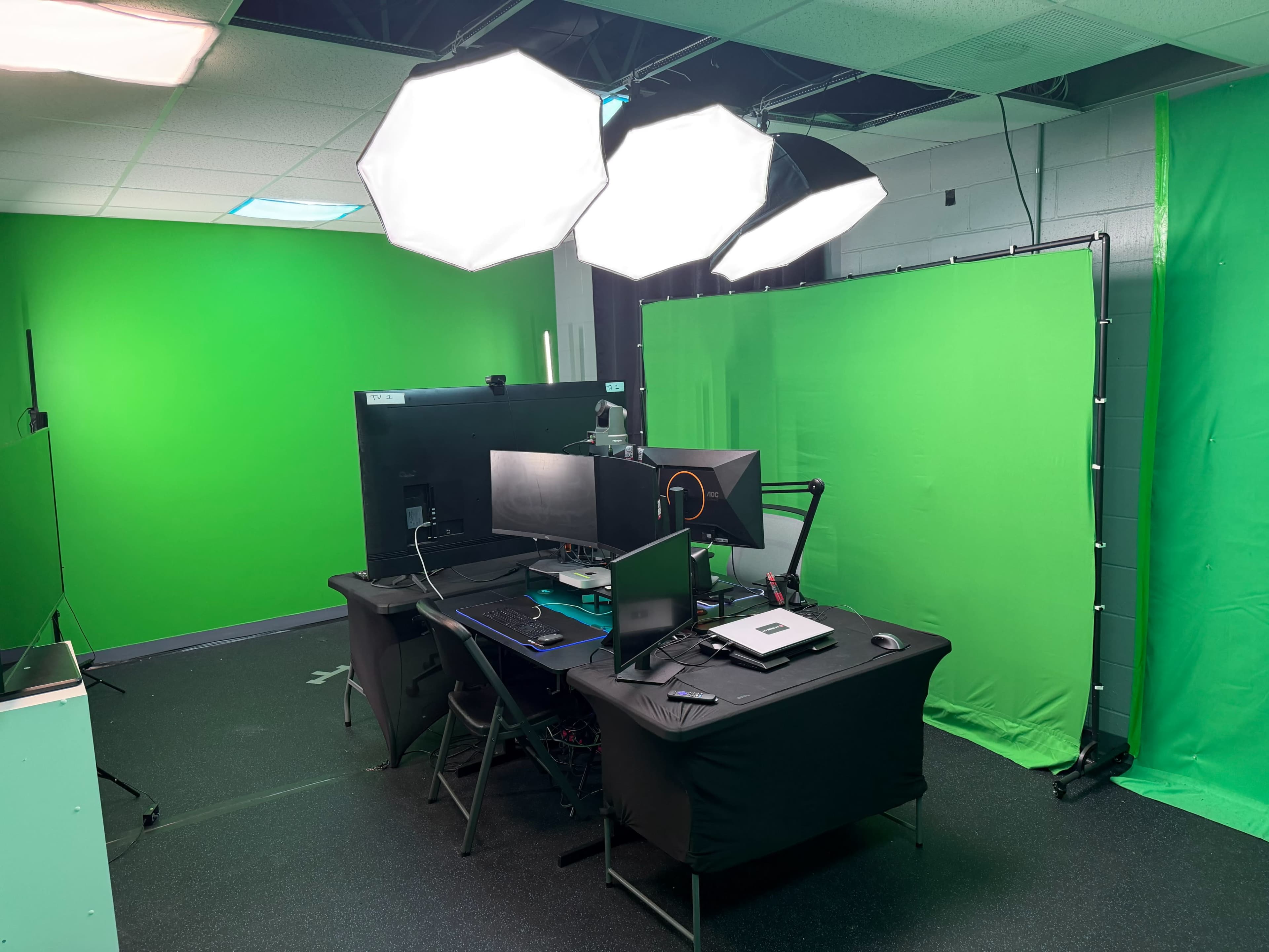 The image shows a studio setup featuring multiple lighting fixtures, a desk with computer monitors, and green backdrop panels for chroma key production.