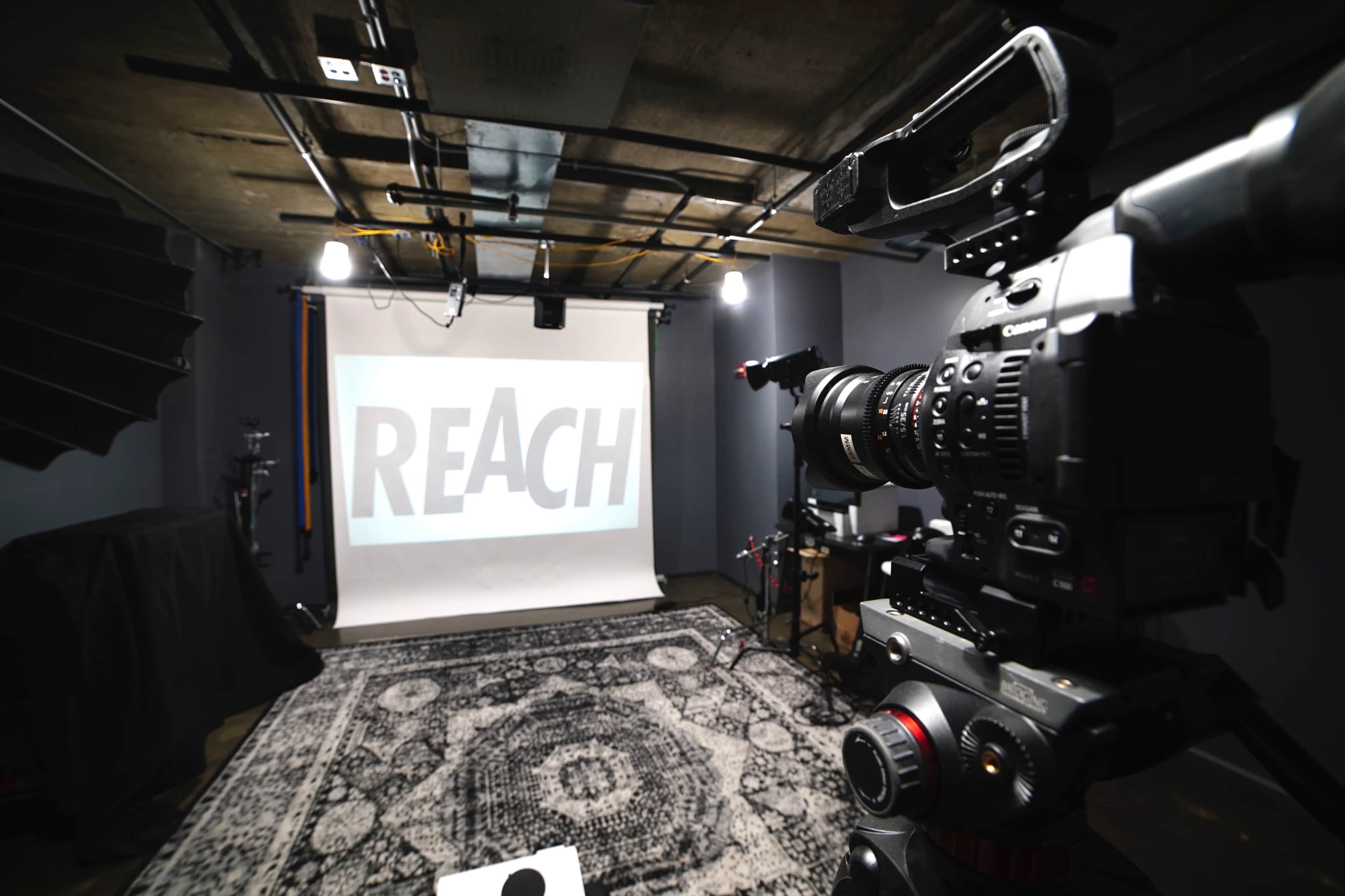 A camera is positioned in front of a projection screen displaying the word "REACH" in a studio setting with dim lighting and a patterned rug.