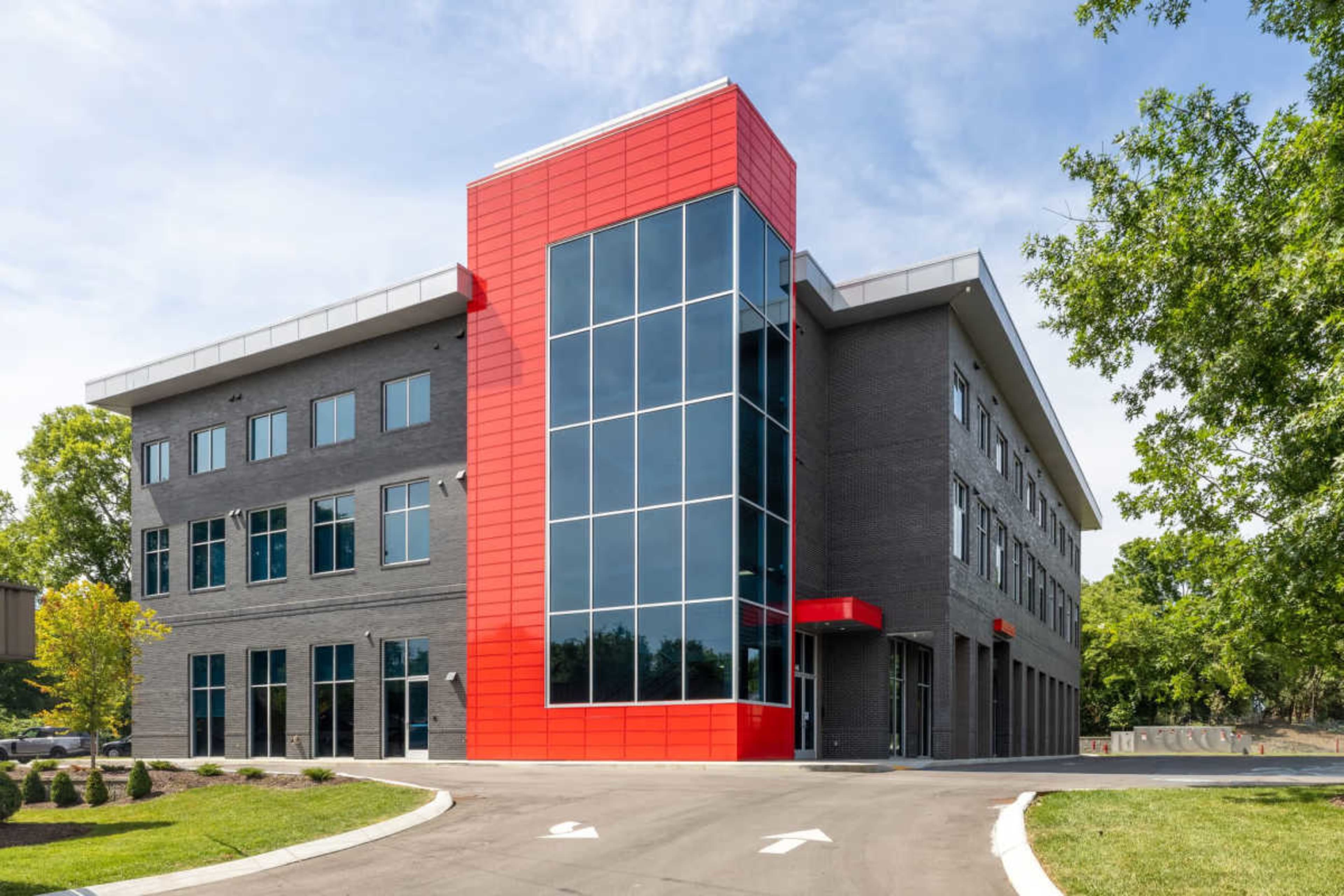 The image shows a modern multi-story building with a prominent red and gray façade, large windows, and a clear sky in the background.