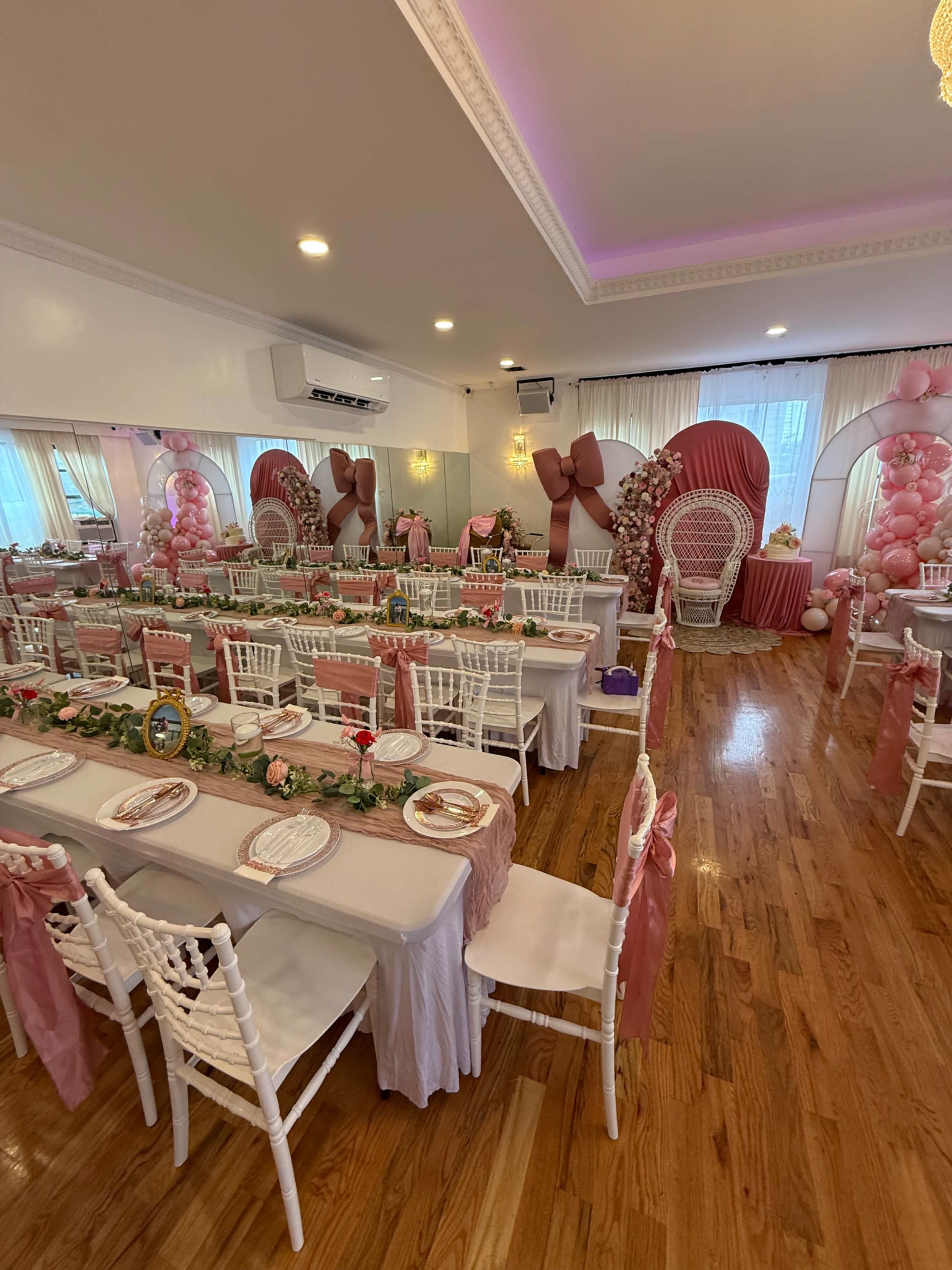 The image shows a decorated banquet hall set up for a celebration, featuring tables with pink and white tablecloths, floral centerpieces, and elegant chairs.