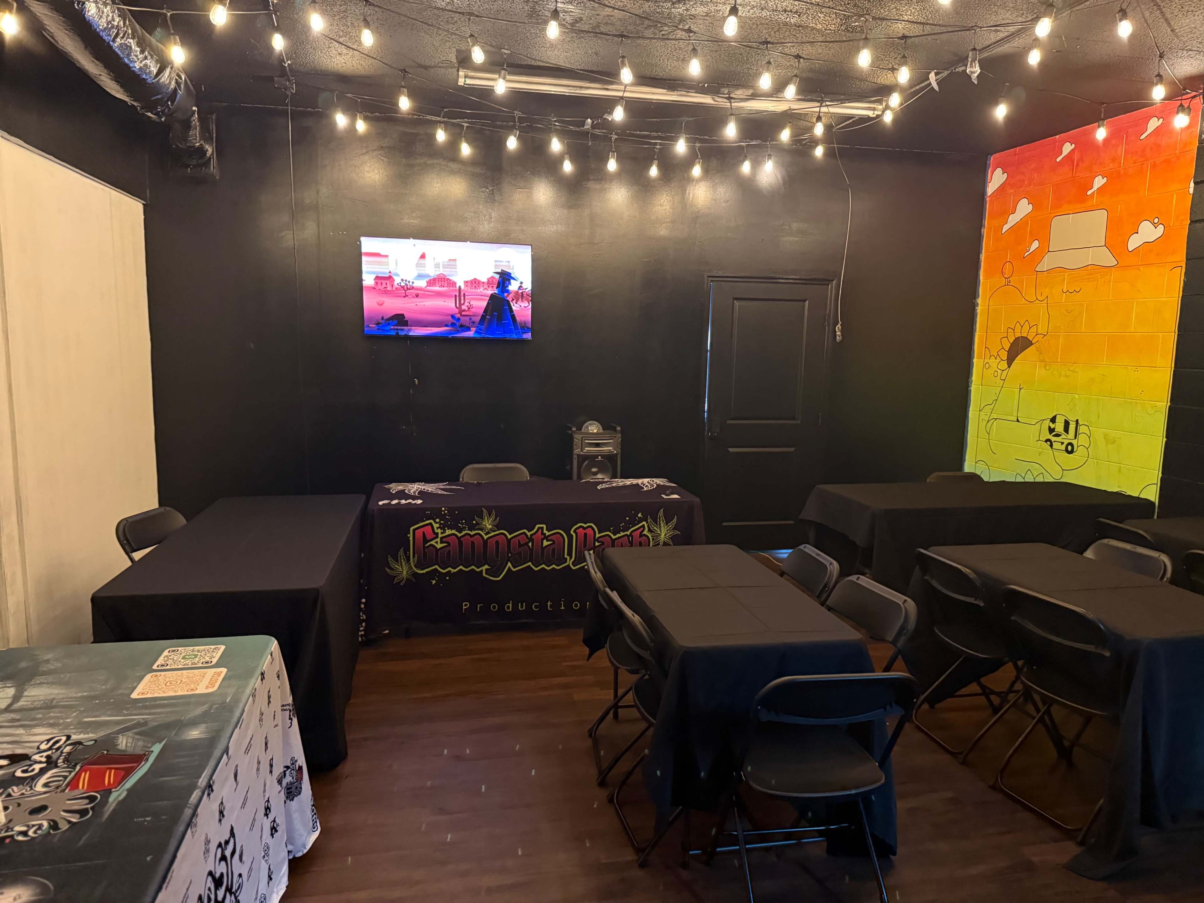A small event space is set up with several tables covered in black cloth, a television mounted on the wall, and string lights overhead.
