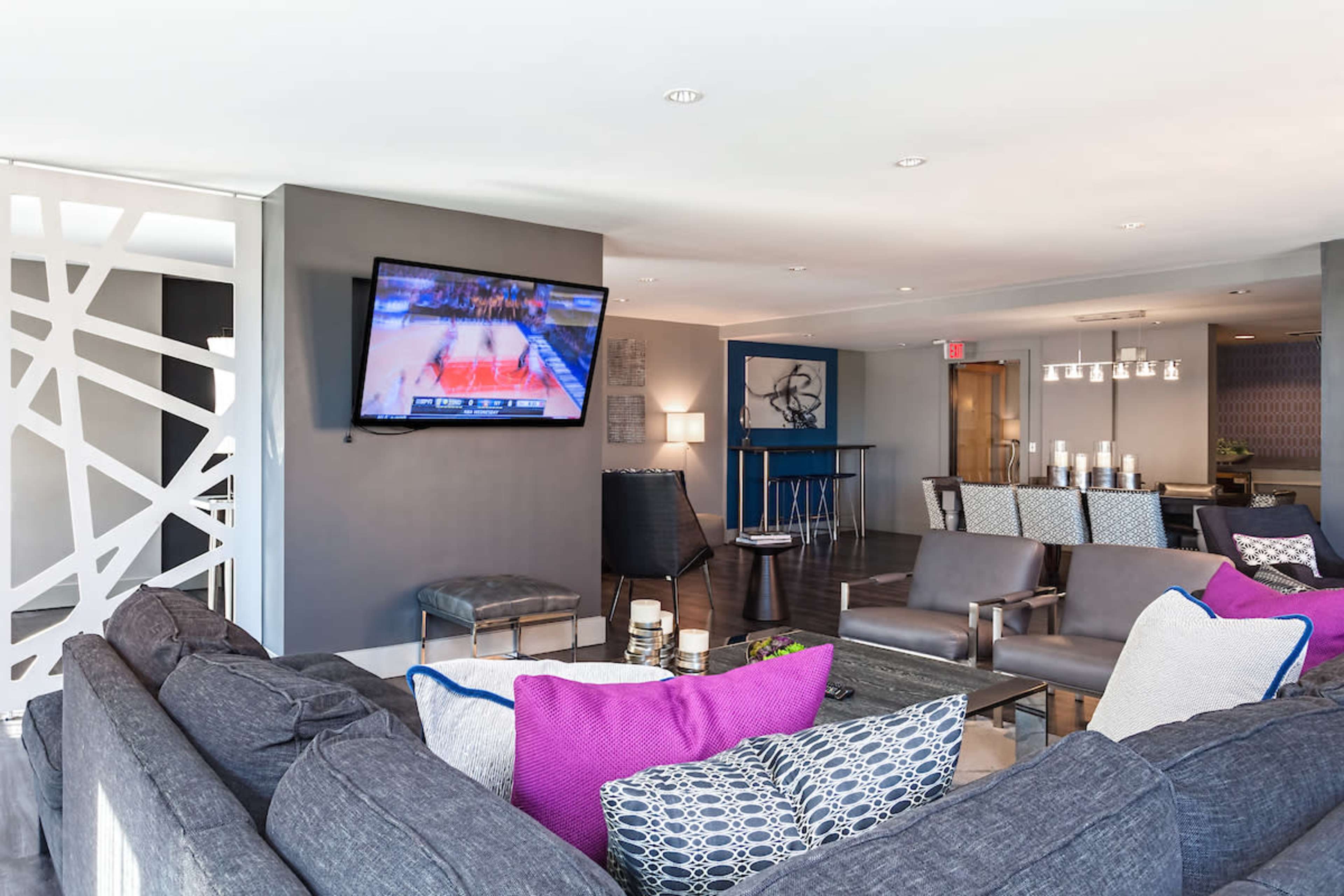 The image shows a modern lounge area with a large television displaying a basketball game, surrounded by comfortable seating and decorative elements.