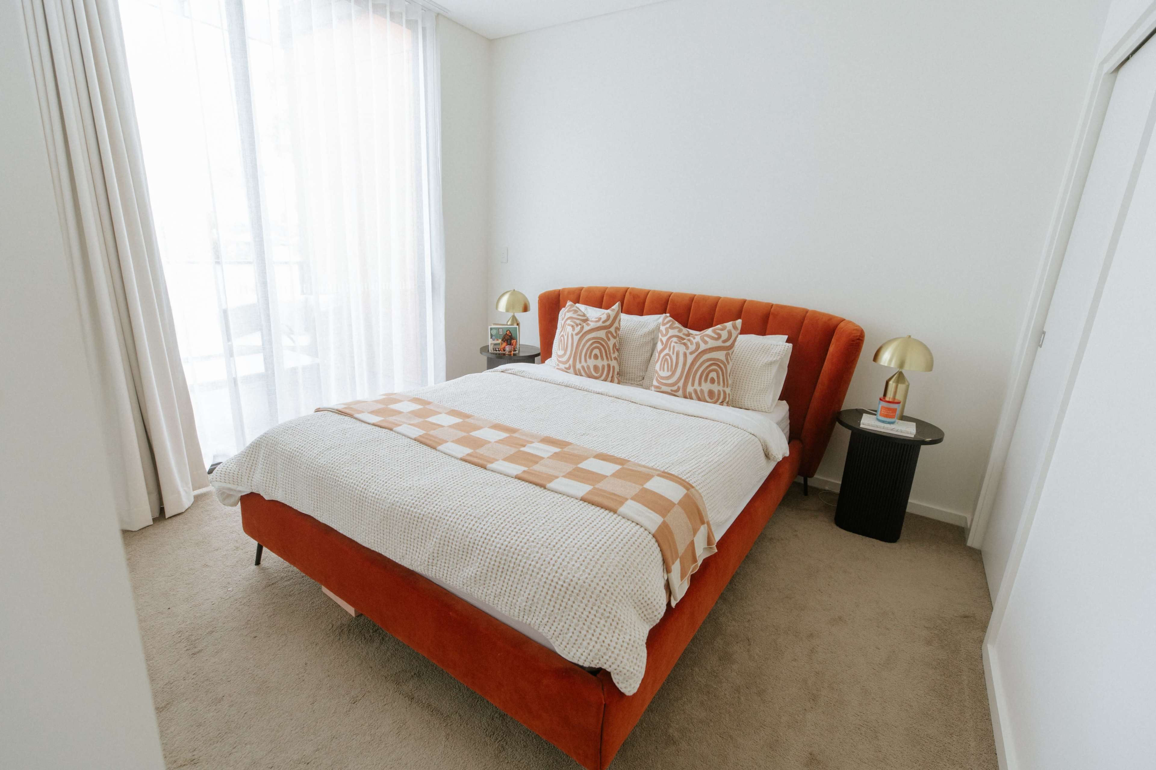 The bedroom features an orange upholstered bed with layered bedding, two patterned pillows, and a bedside table with a lamp.