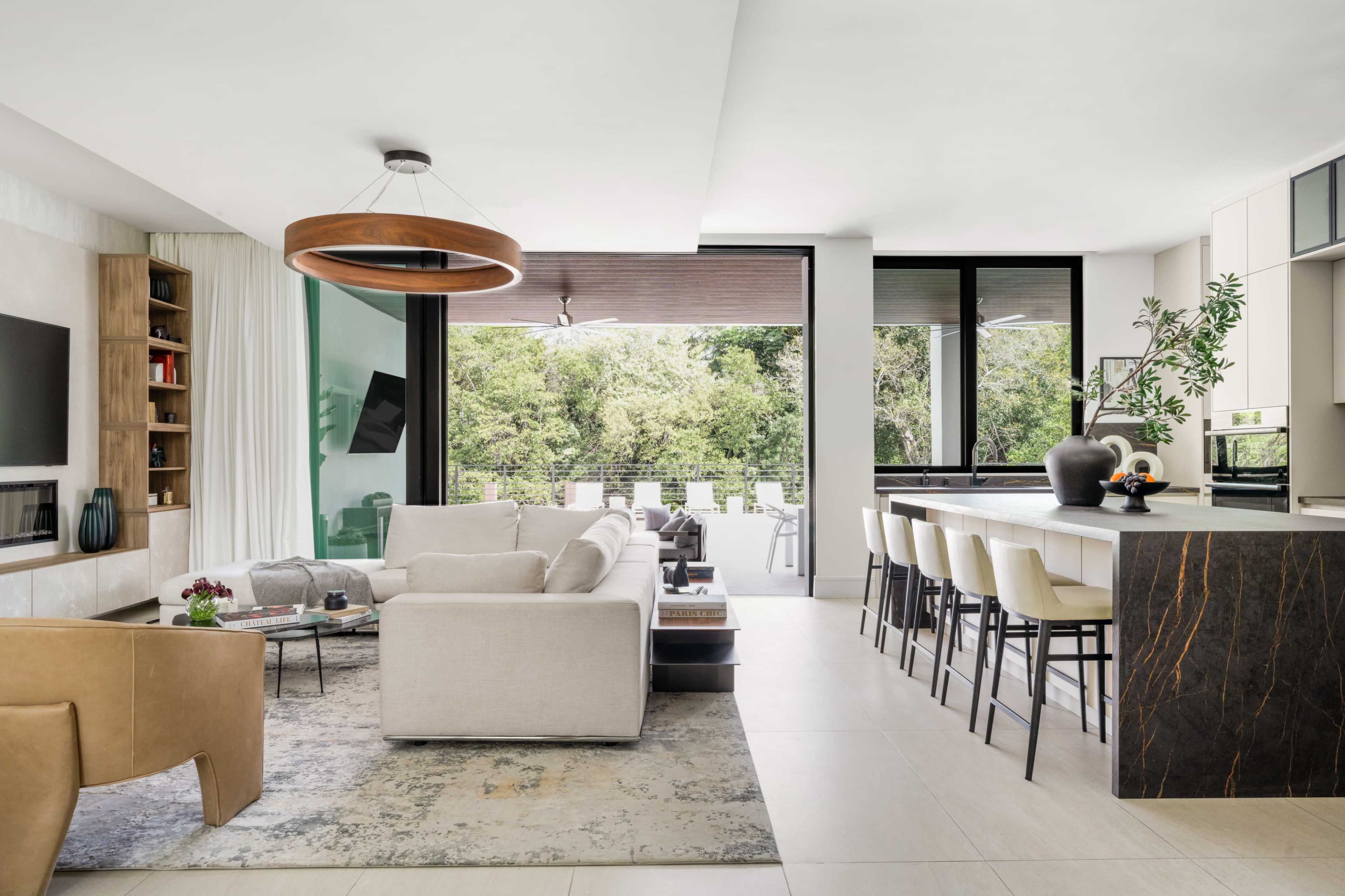 A modern living space featuring a light-colored sofa, an open kitchen with bar stools, and large windows overlooking a green outdoor area.