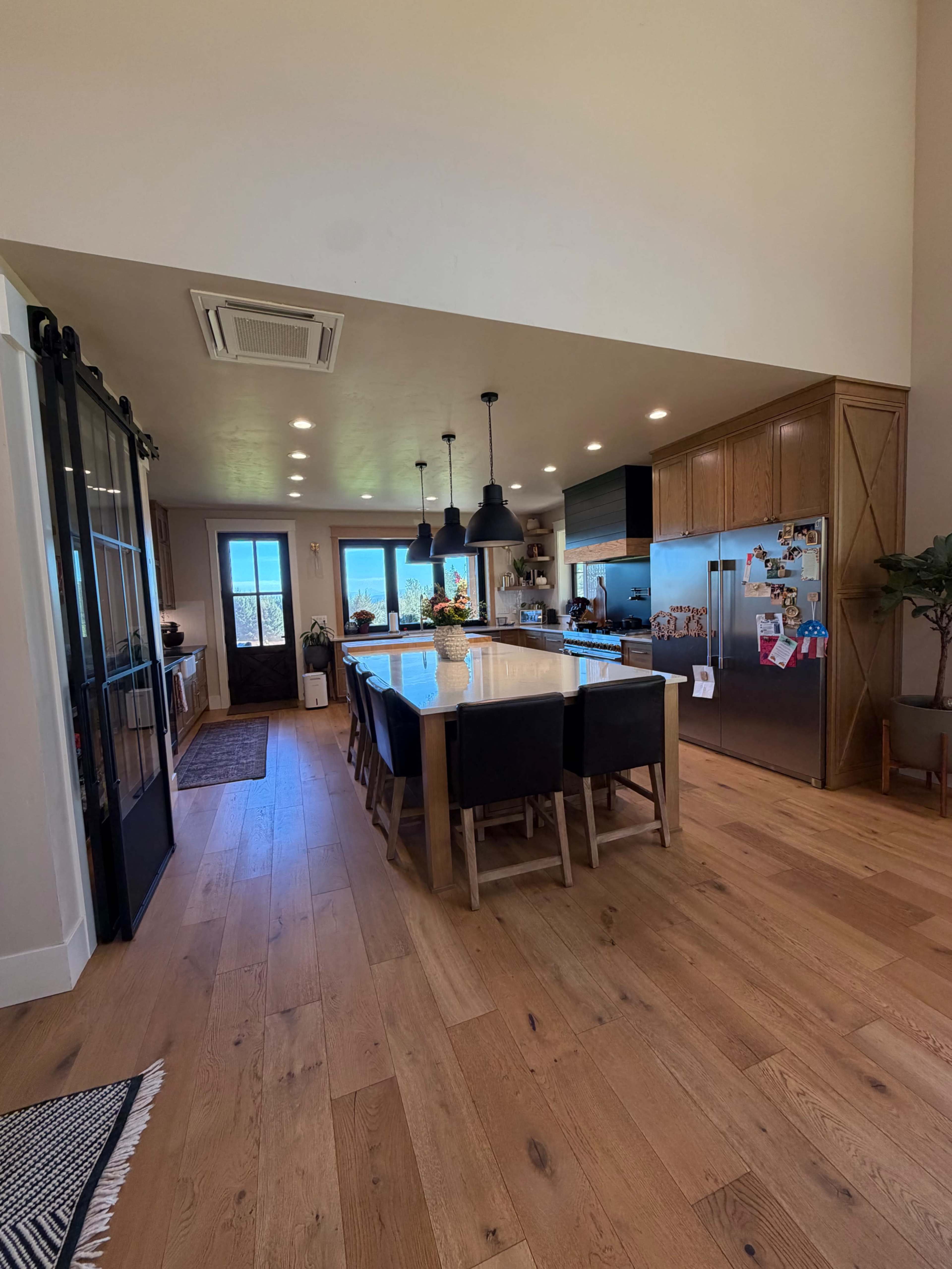 The image shows a modern kitchen with a large island, wooden cabinetry, and several windows allowing natural light to enter.
