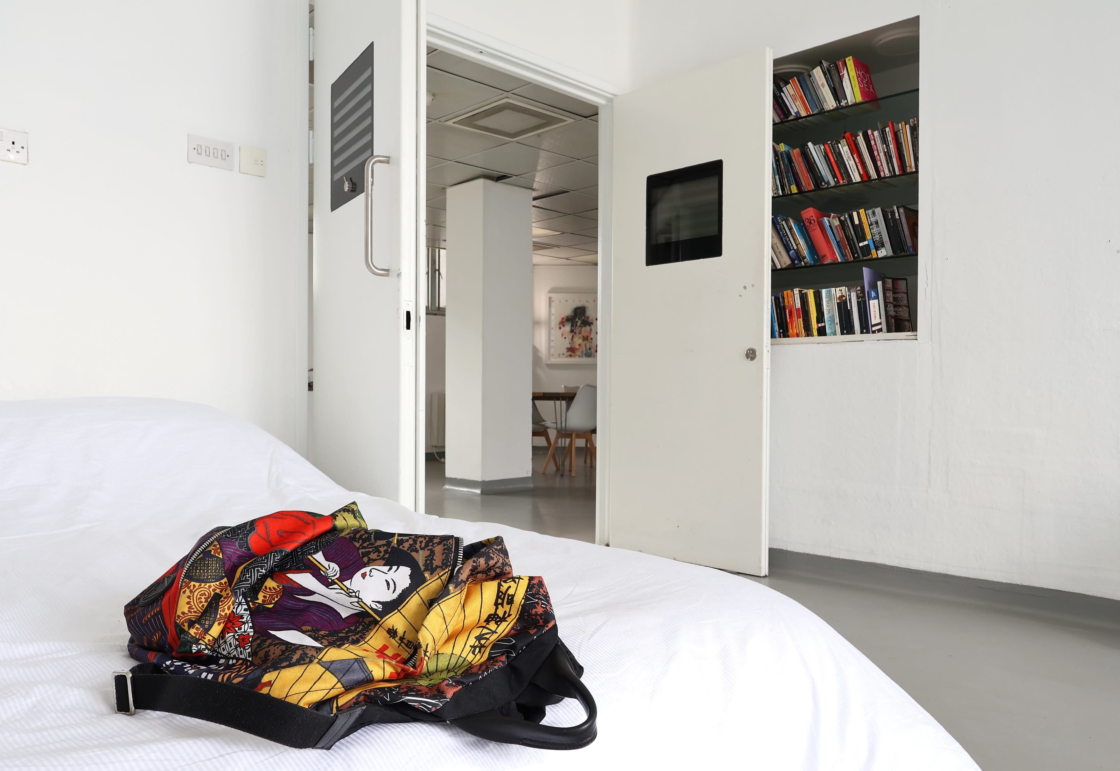 A colorful bag lies on a bed in a minimally furnished room with an open door leading to a bookshelf and another area.